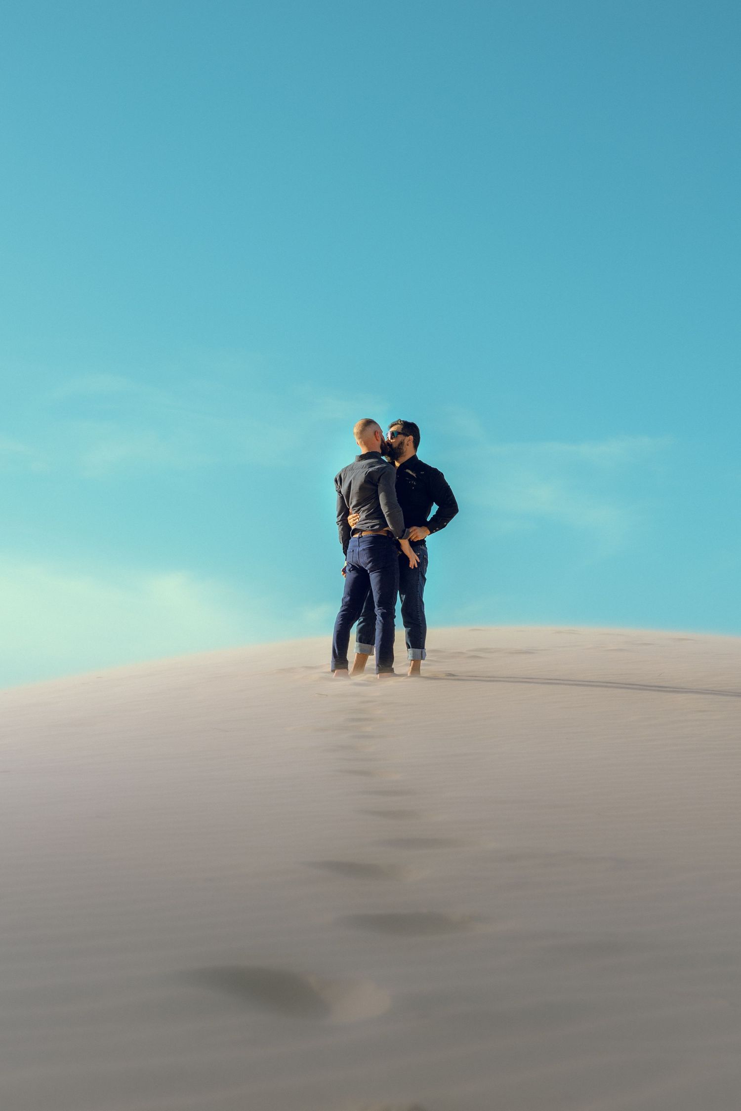 Couple walking together on sand dunes against bright blue sky.