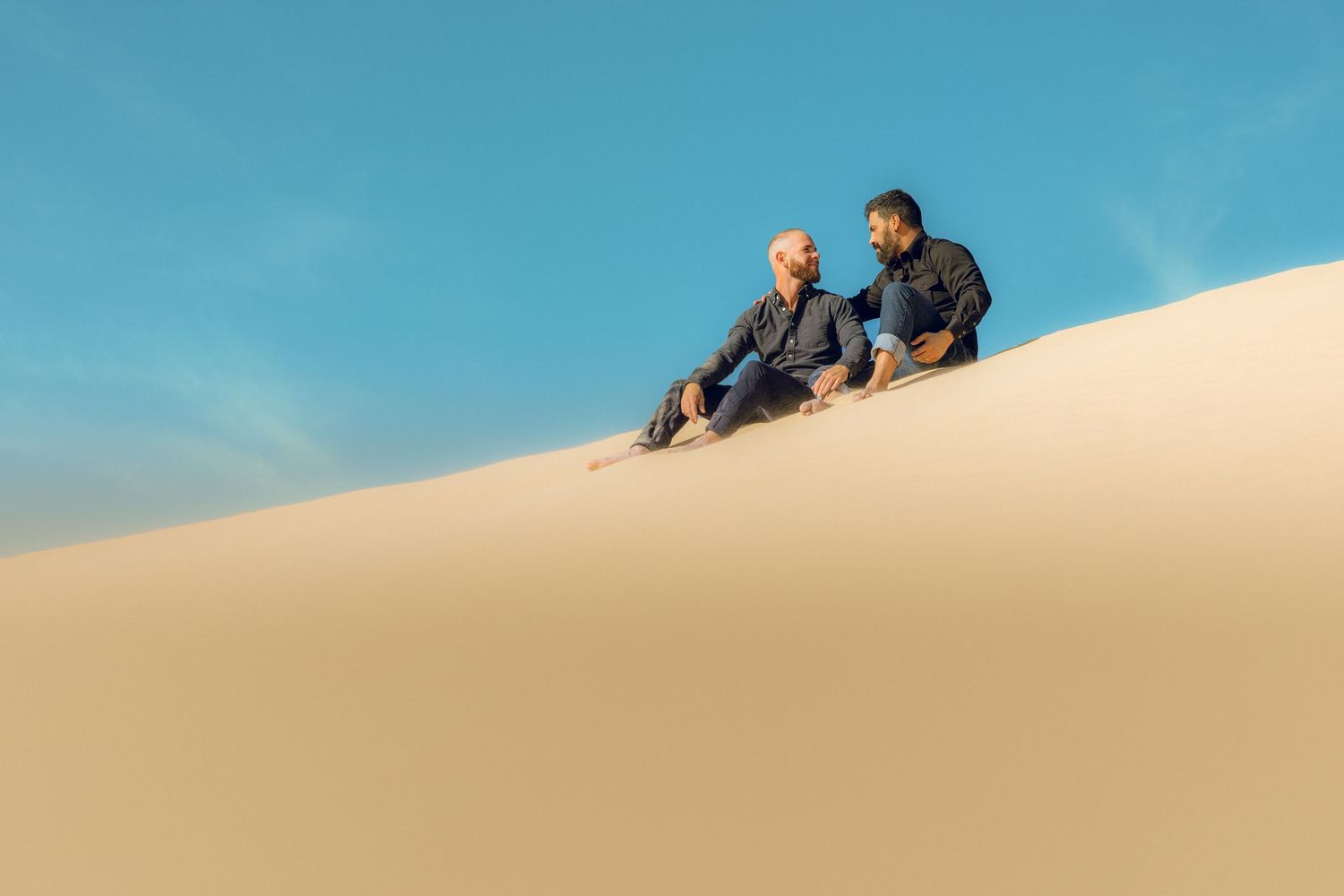 Two people sitting together on sandy ridge against bright blue desert sky.