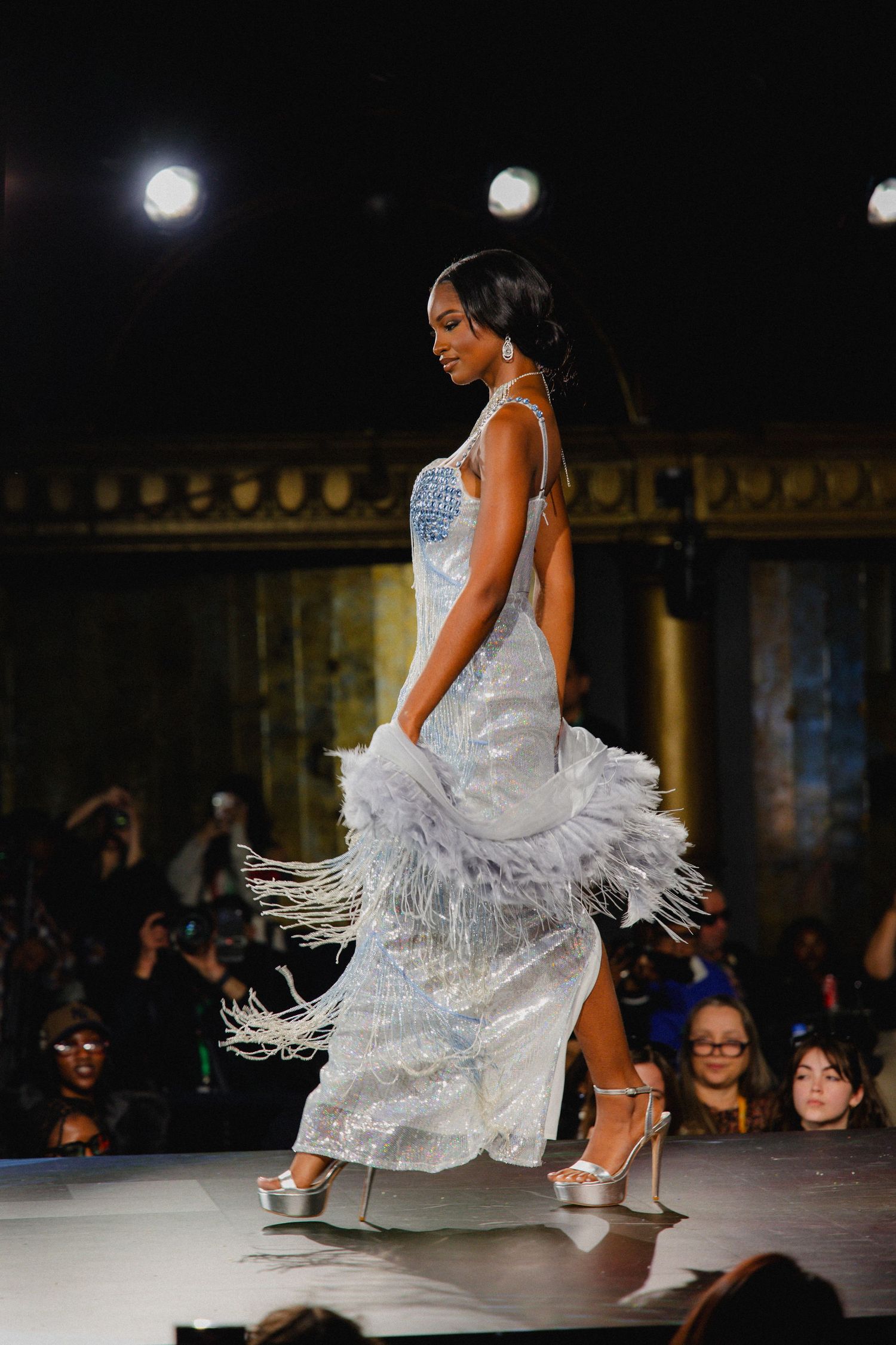 Fashion model in white feathered dress twirls dramatically on runway during fashion show.