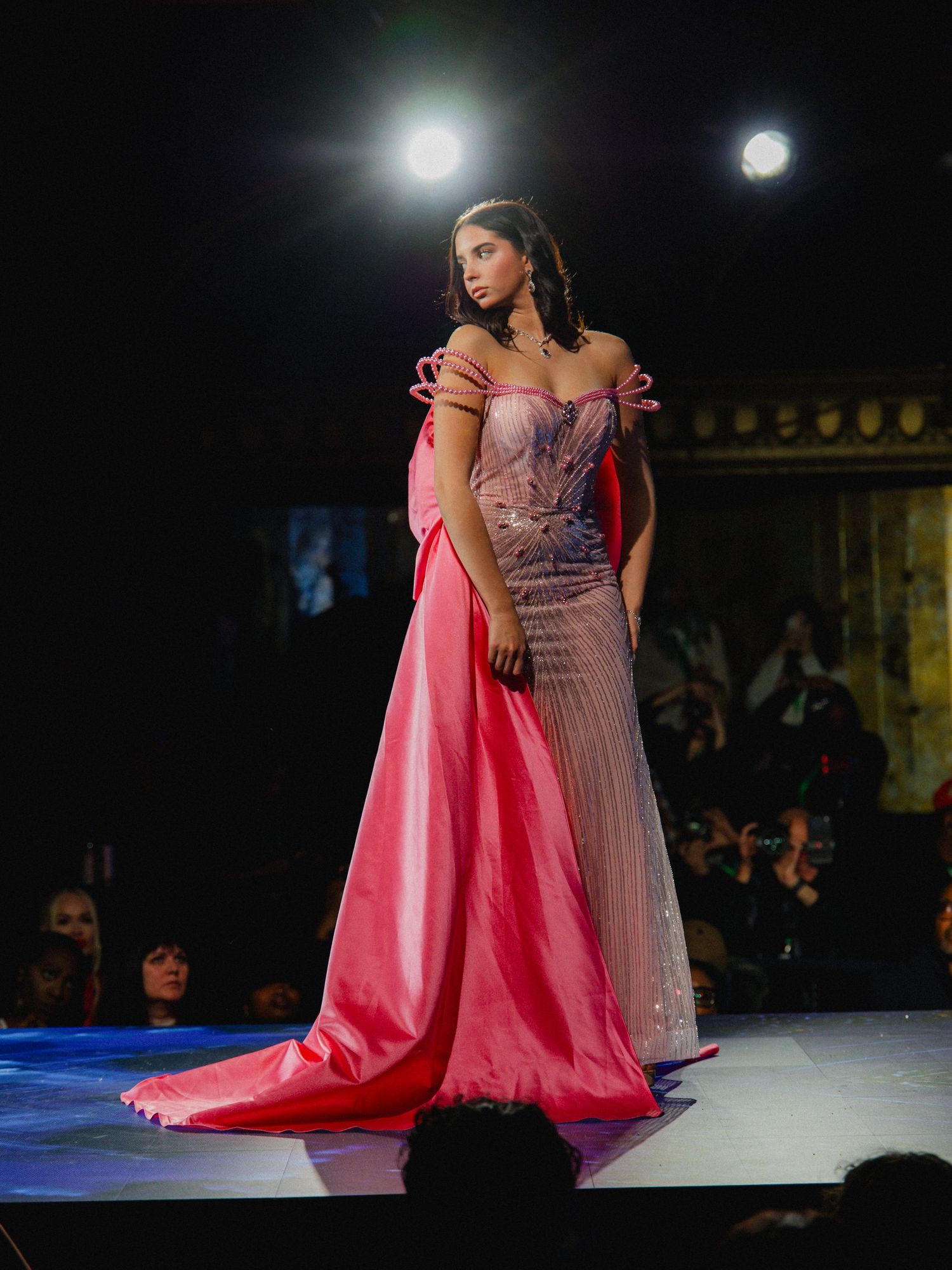 Model walks runway in stunning pink evening gown with dramatic cape and fitted bodice at fashion show.