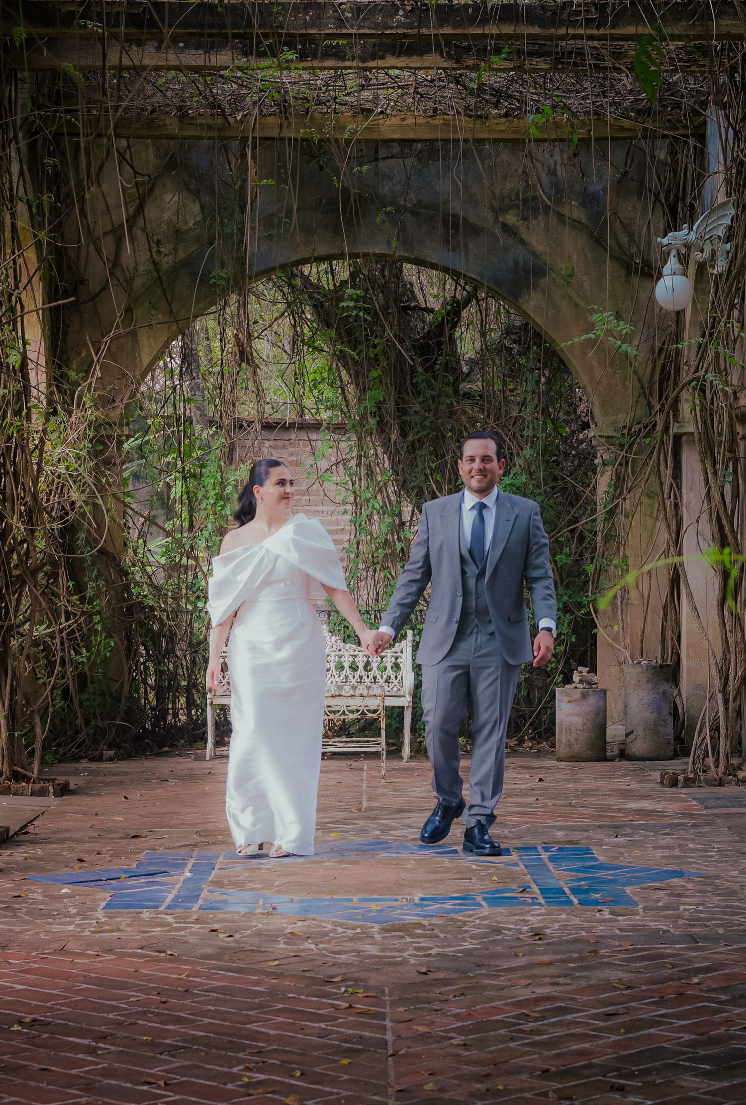 Couple walking hand in hand under a vine-covered archway in a garden setting.