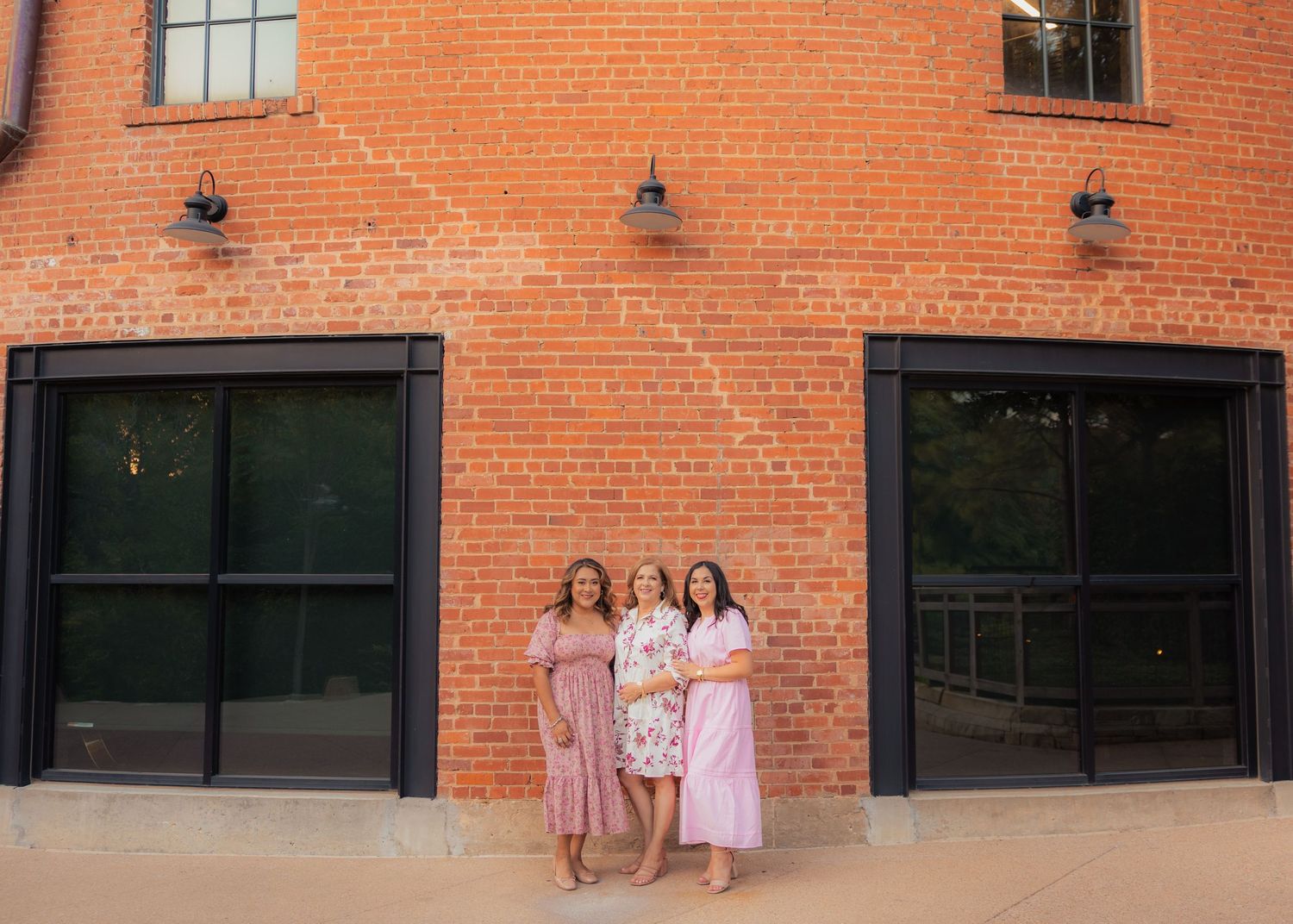 Three people in pink outfits standing against a brick wall with windows.