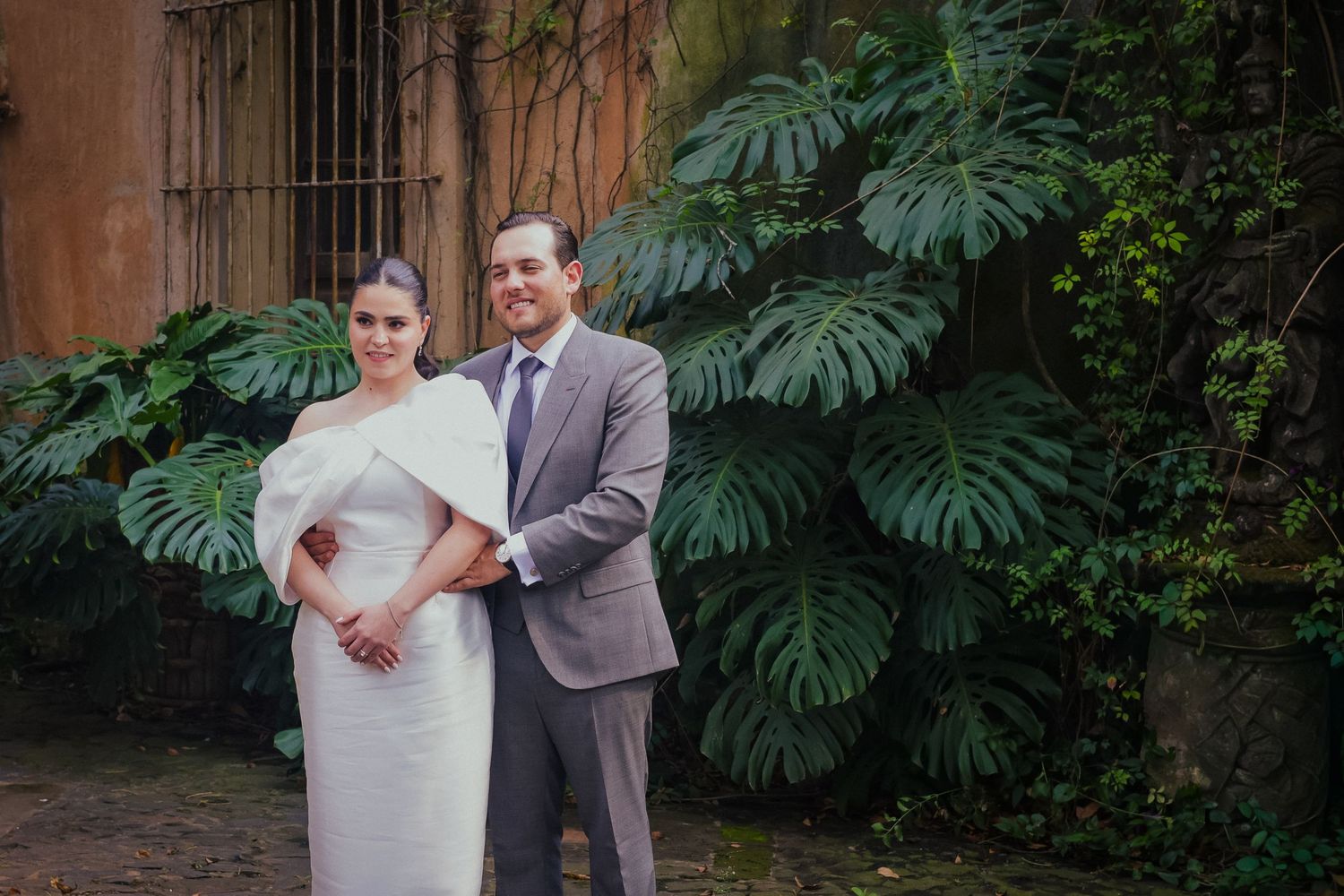 Wedding couple poses in formal attire against lush tropical garden backdrop.
