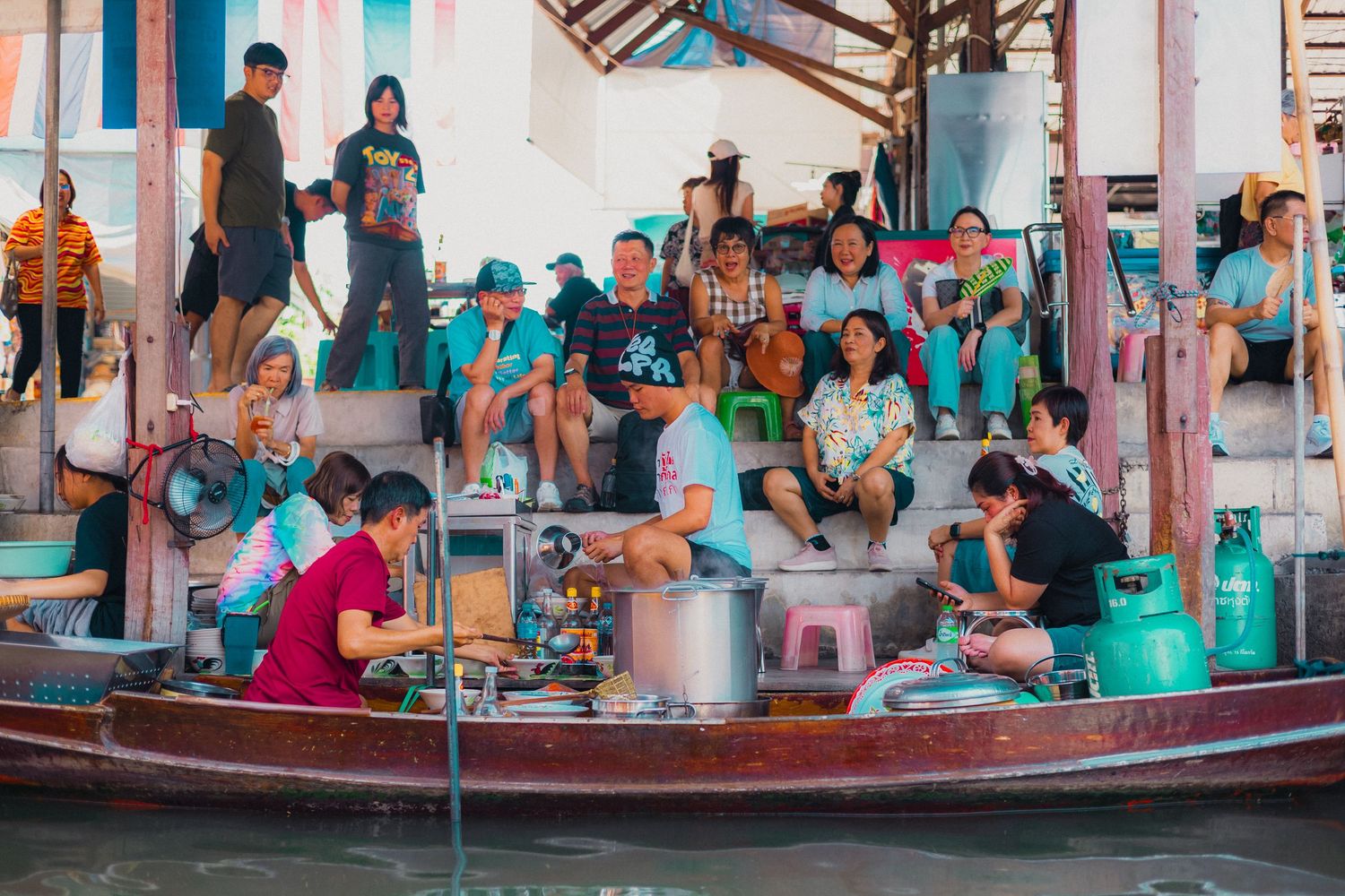 Busy floating market with vendors cooking on boats and tourists watching from a wooden dock in Thailand.