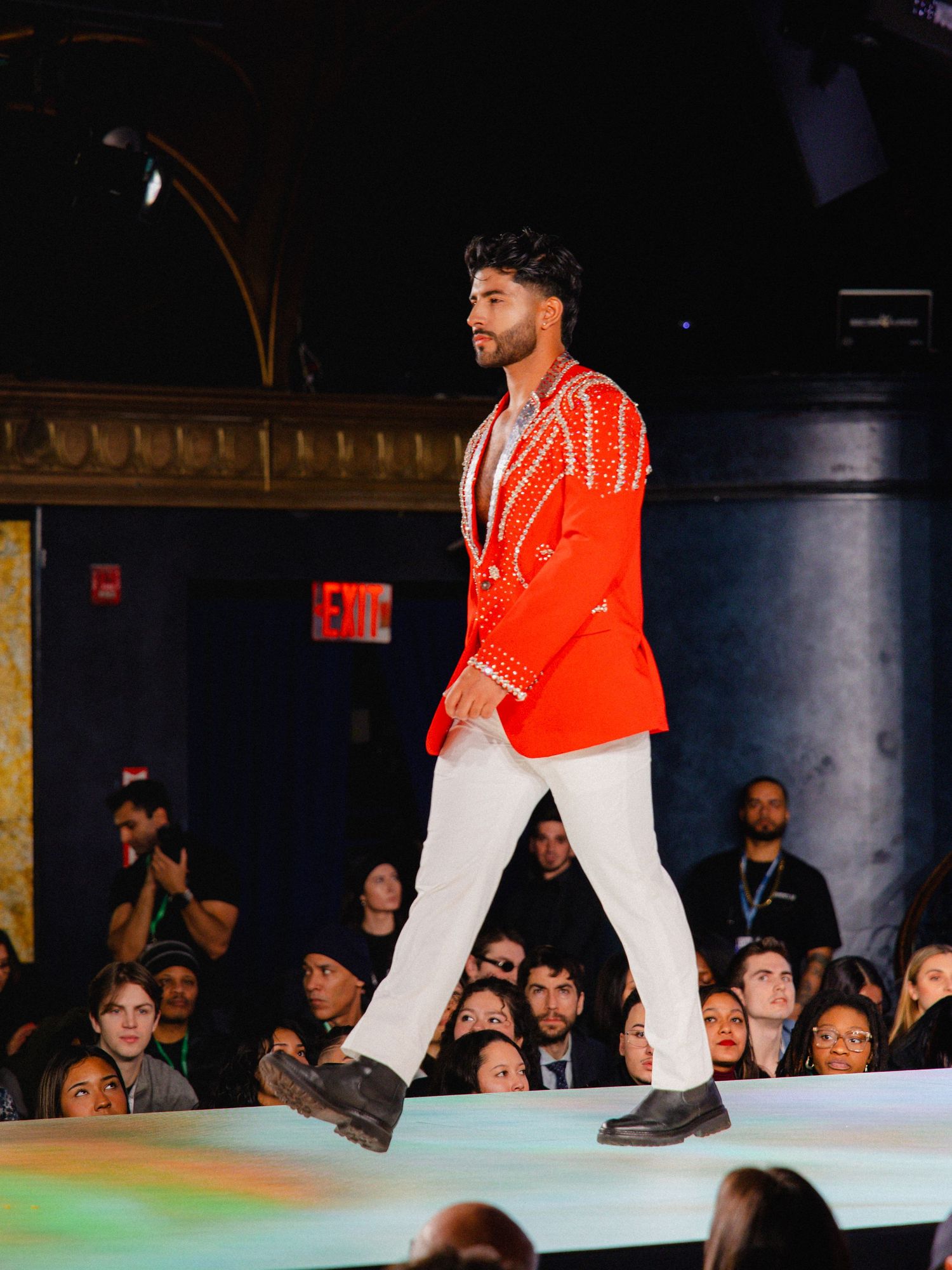 Fashion model wearing orange and white outfit walks down illuminated runway during fashion show event.