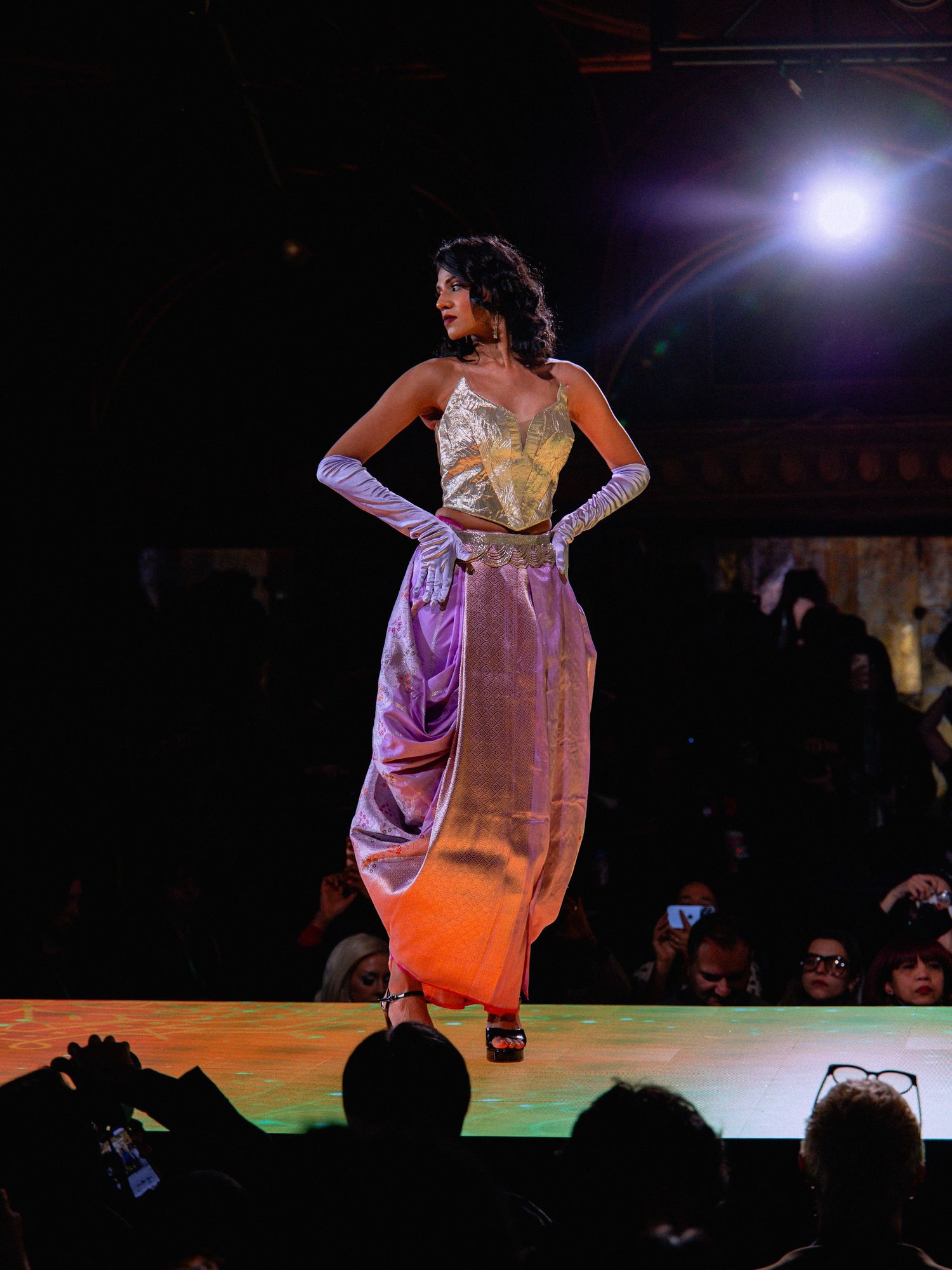 Model walks runway in flowing lavender evening gown under dramatic stage lighting at fashion show.