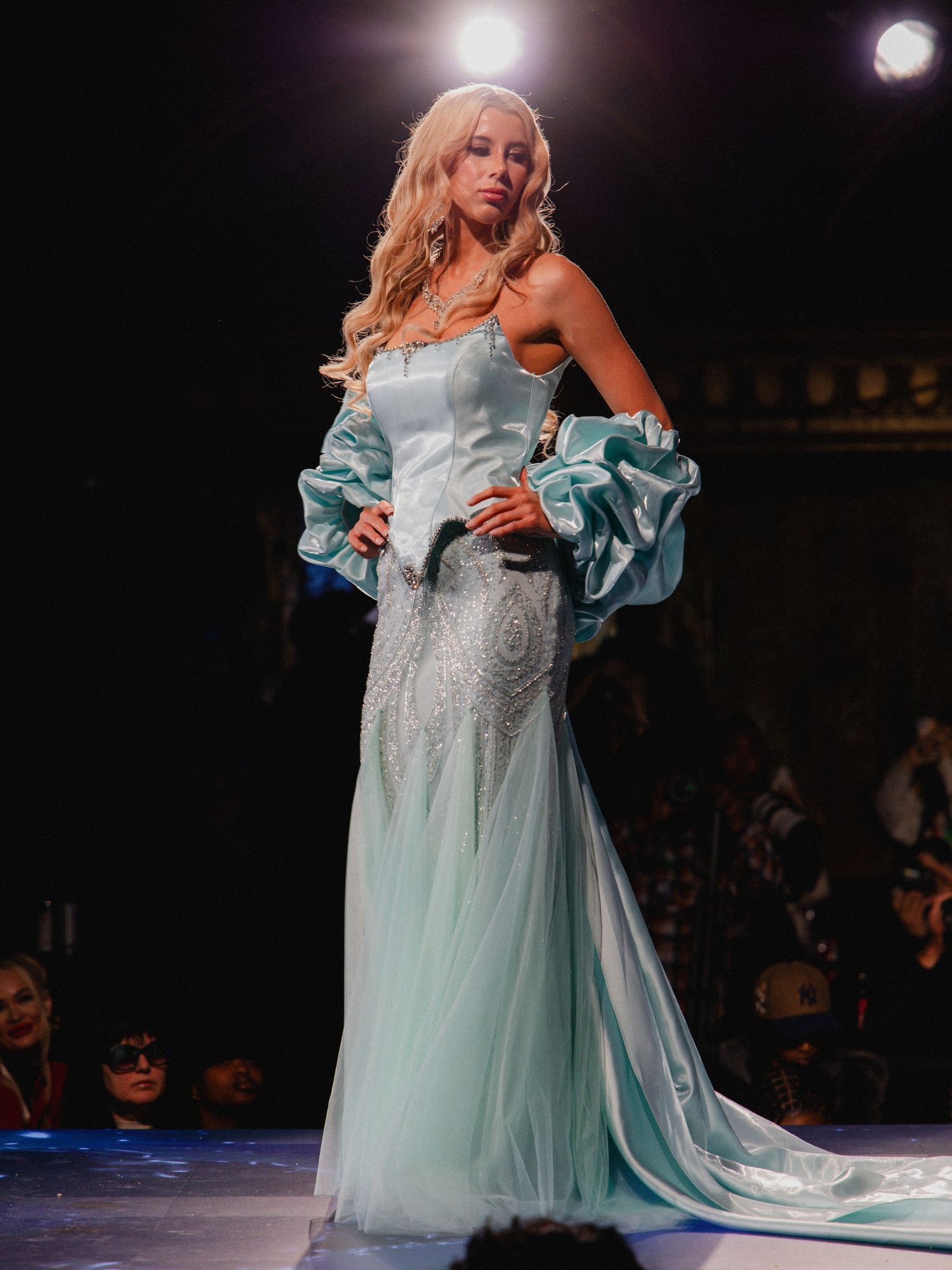A performer in a flowing light blue gown with off-shoulder sleeves twirls gracefully on a dark stage under spotlights.