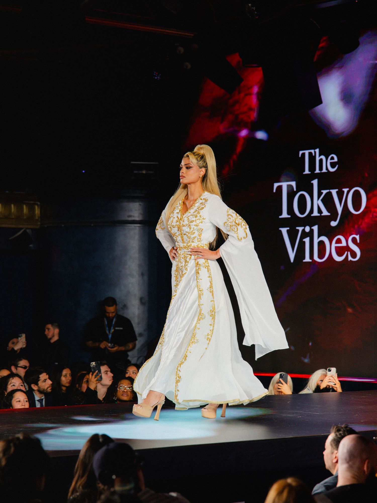 A model walks down a dark runway in a flowing white dress with dramatic sleeves during a fashion show.