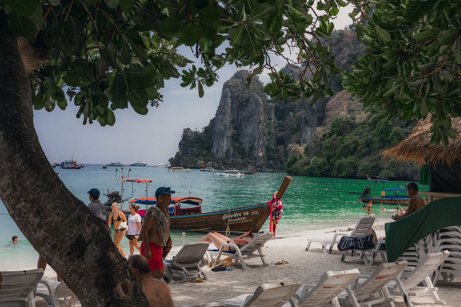 Tourists relax on white sand beach with turquoise water, longtail boats, and dramatic limestone cliffs in Koh Phi Phi, Thailand.