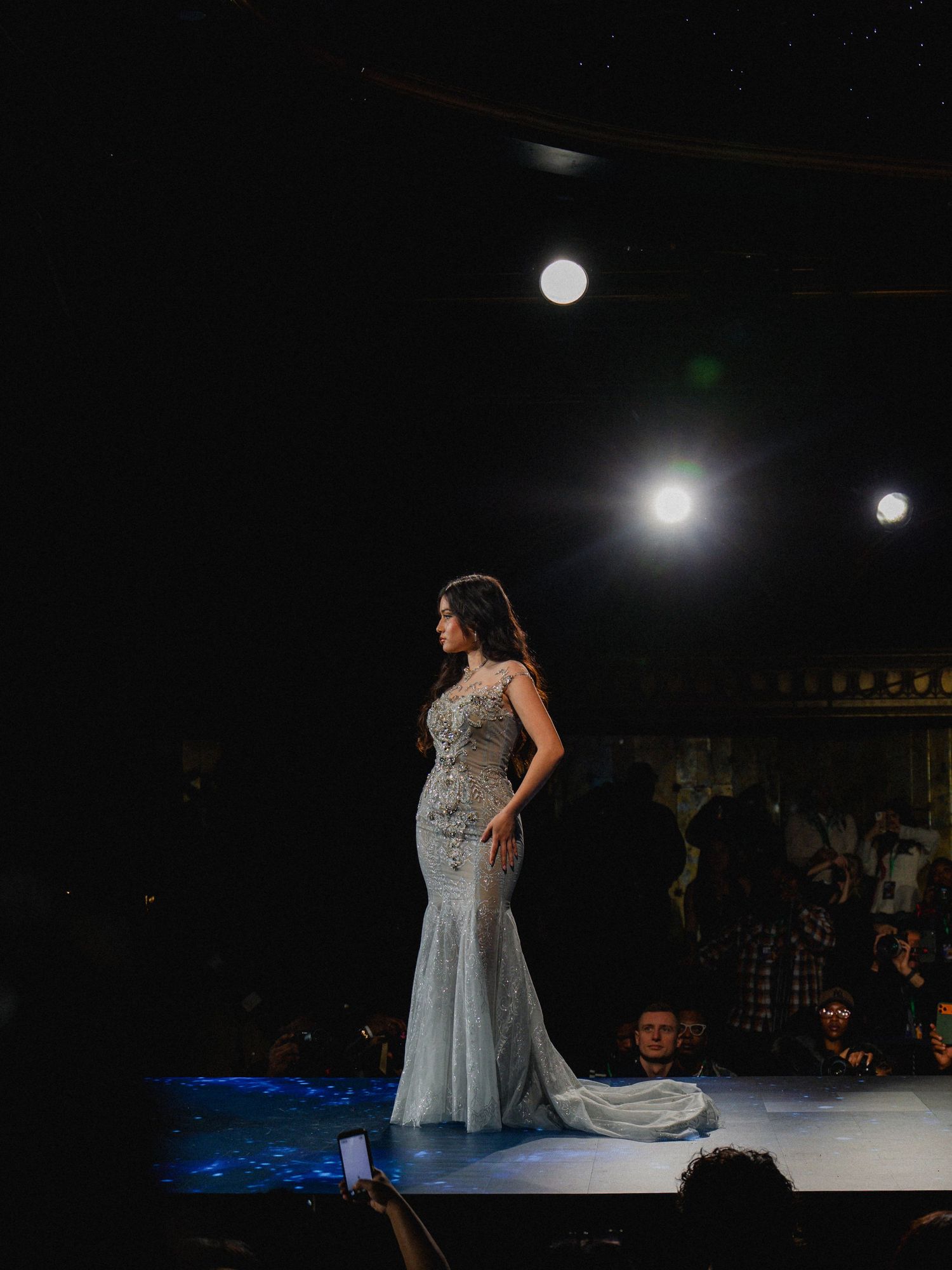 A model in a flowing silver gown poses dramatically on a dark stage during a fashion show.