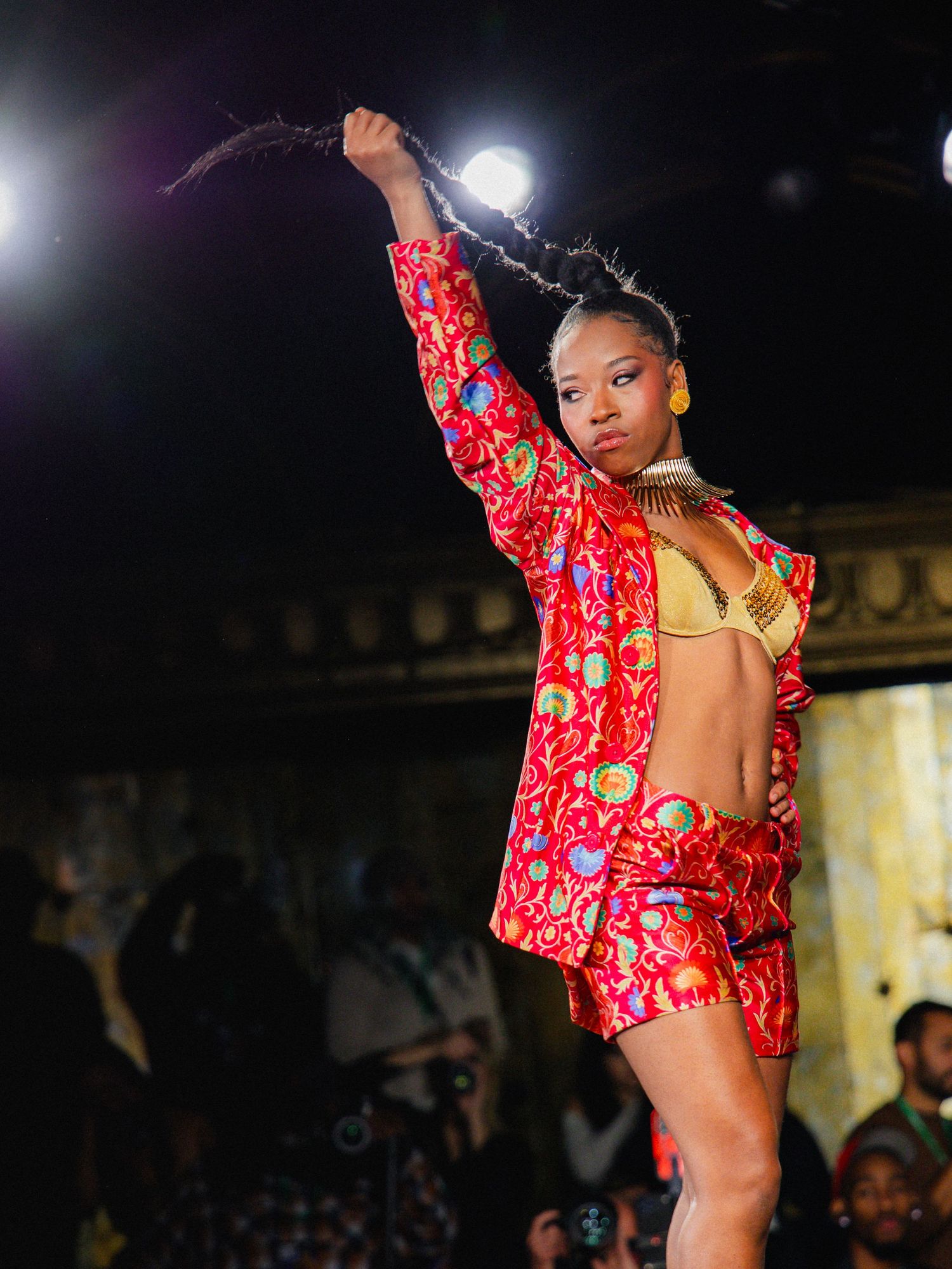 Model walks runway in red floral printed outfit during fashion show with dramatic lighting and dark background.