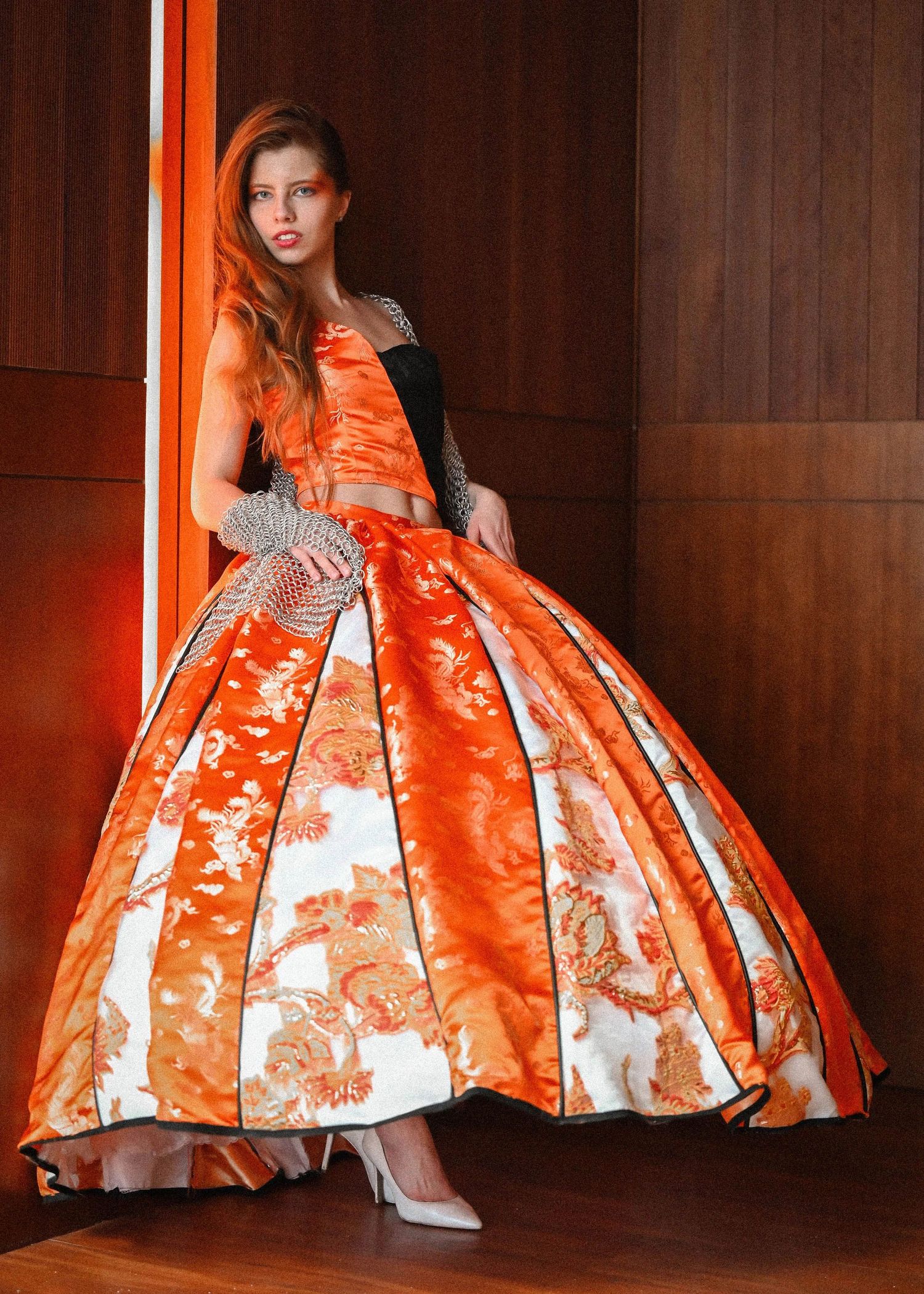 Model wearing dramatic orange ball gown with floral pattern in wood-paneled room.