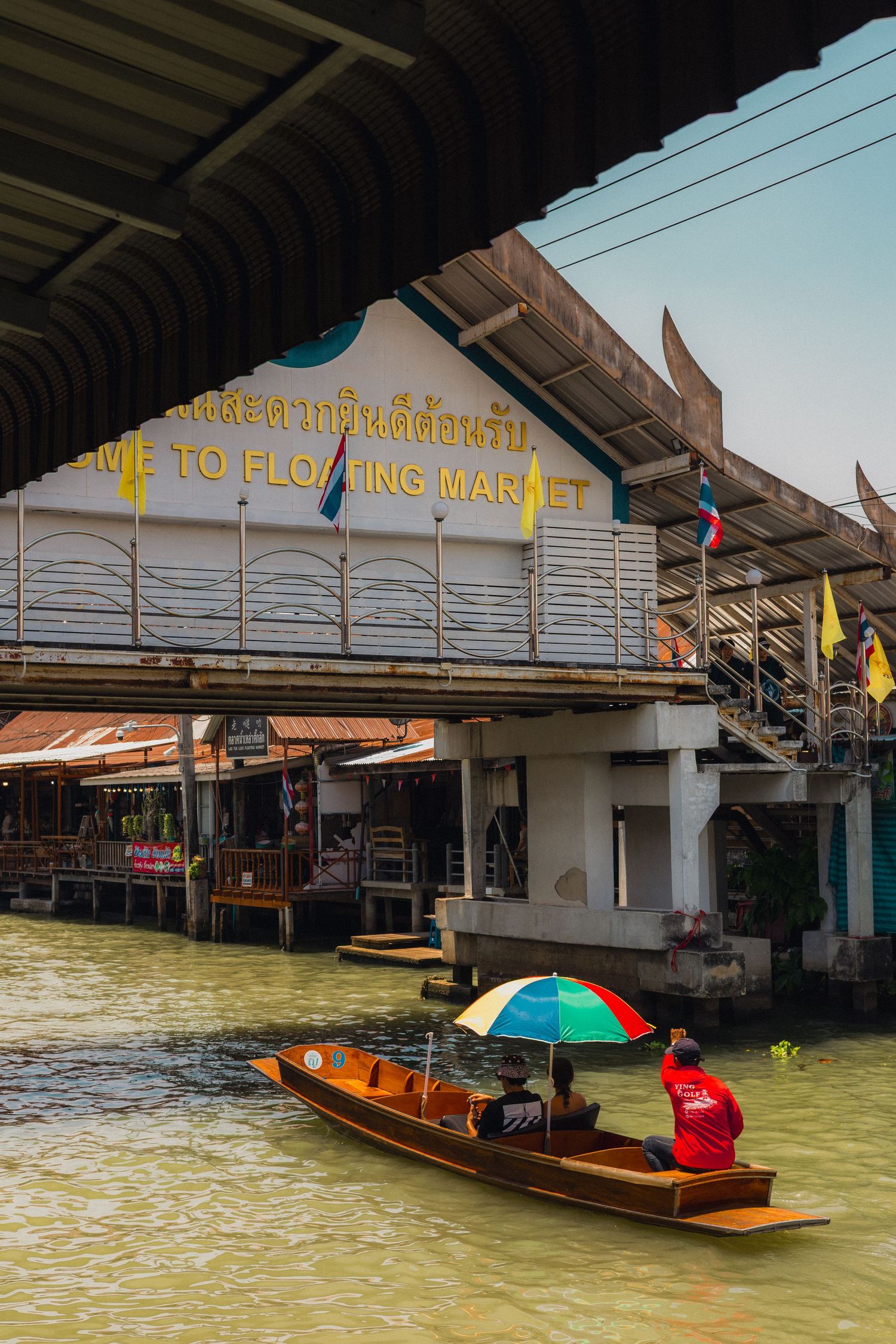 Wooden boat with colorful umbrella gliding along a canal at a traditional Thai floating market with welcome sign.