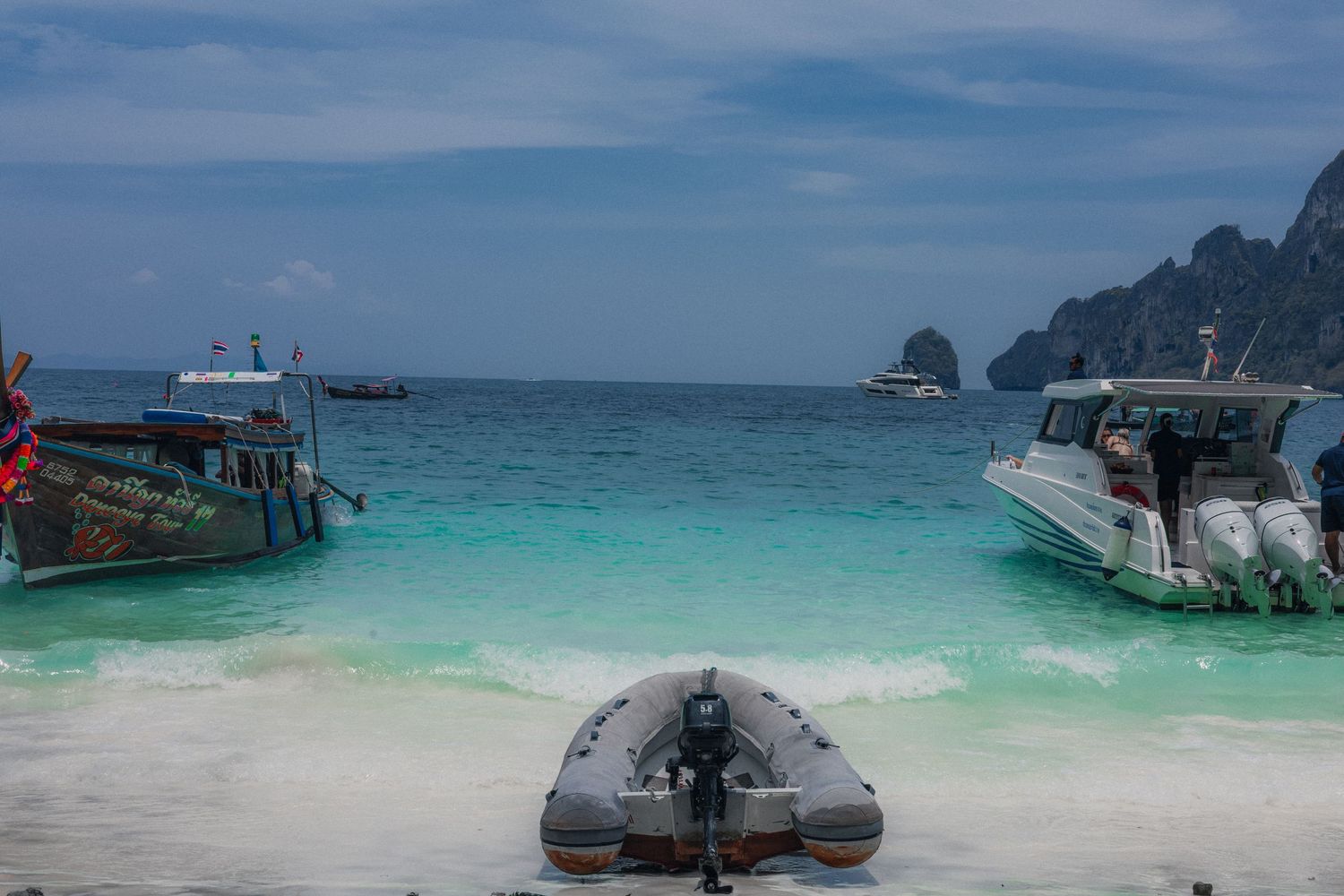 Boats anchored on a turquoise beach in Koh Phi Phi, Thailand, with limestone cliffs and cloudy skies in the background.