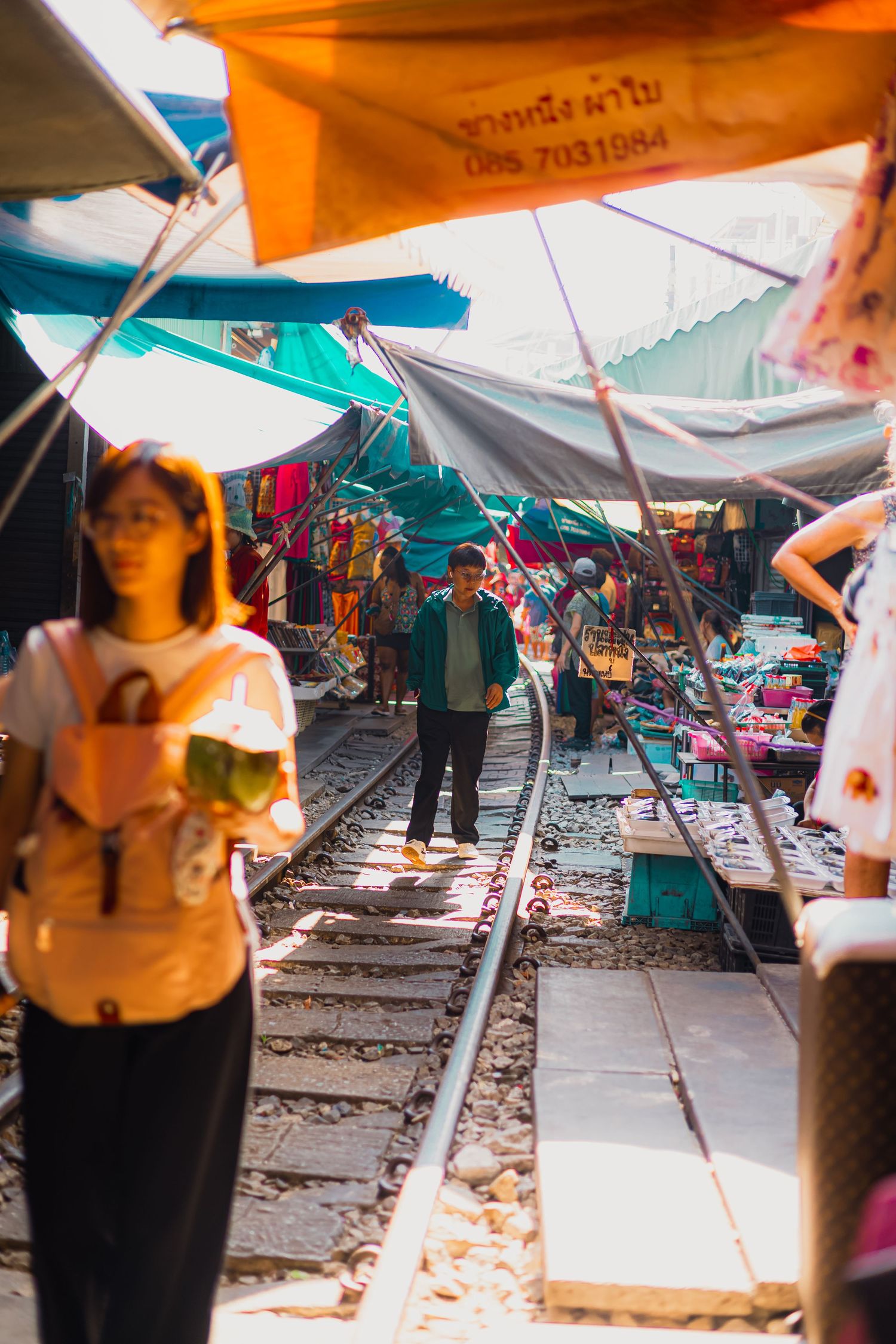 Tourists walk along railway tracks through a vibrant outdoor market with colorful canopies in Thailand.