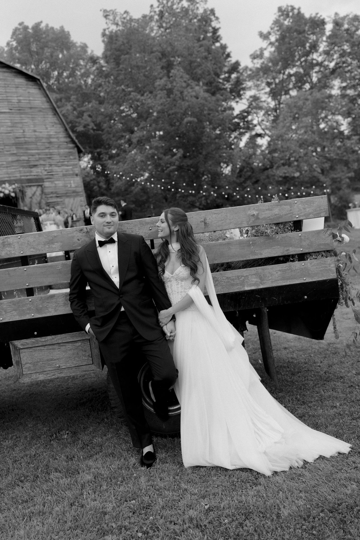 Black and white photo of a bride and groom posing by a rustic wooden wagon at a barn wedding venue.