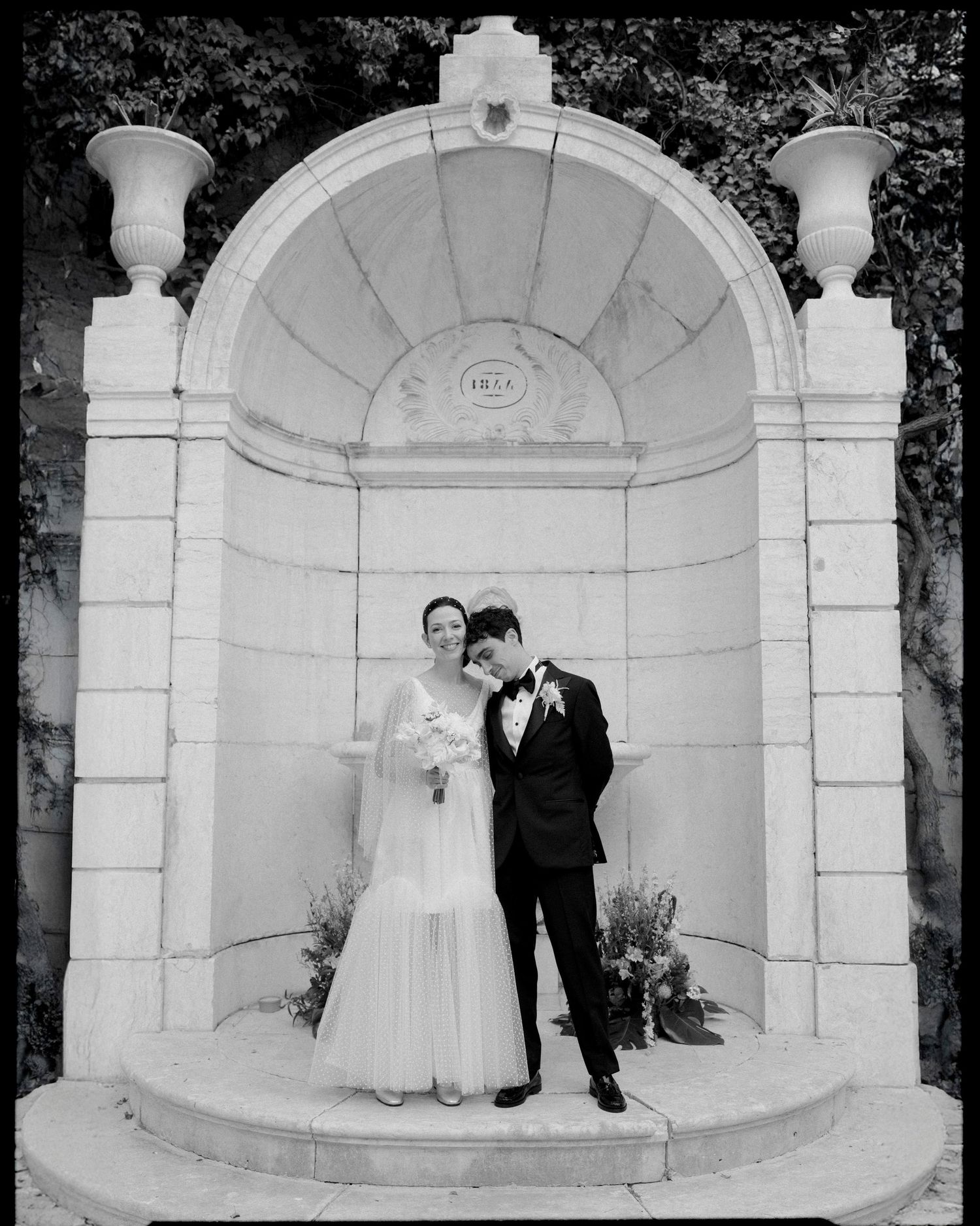 Bride and groom posing under stone arch with decorative urns during wedding portrait
