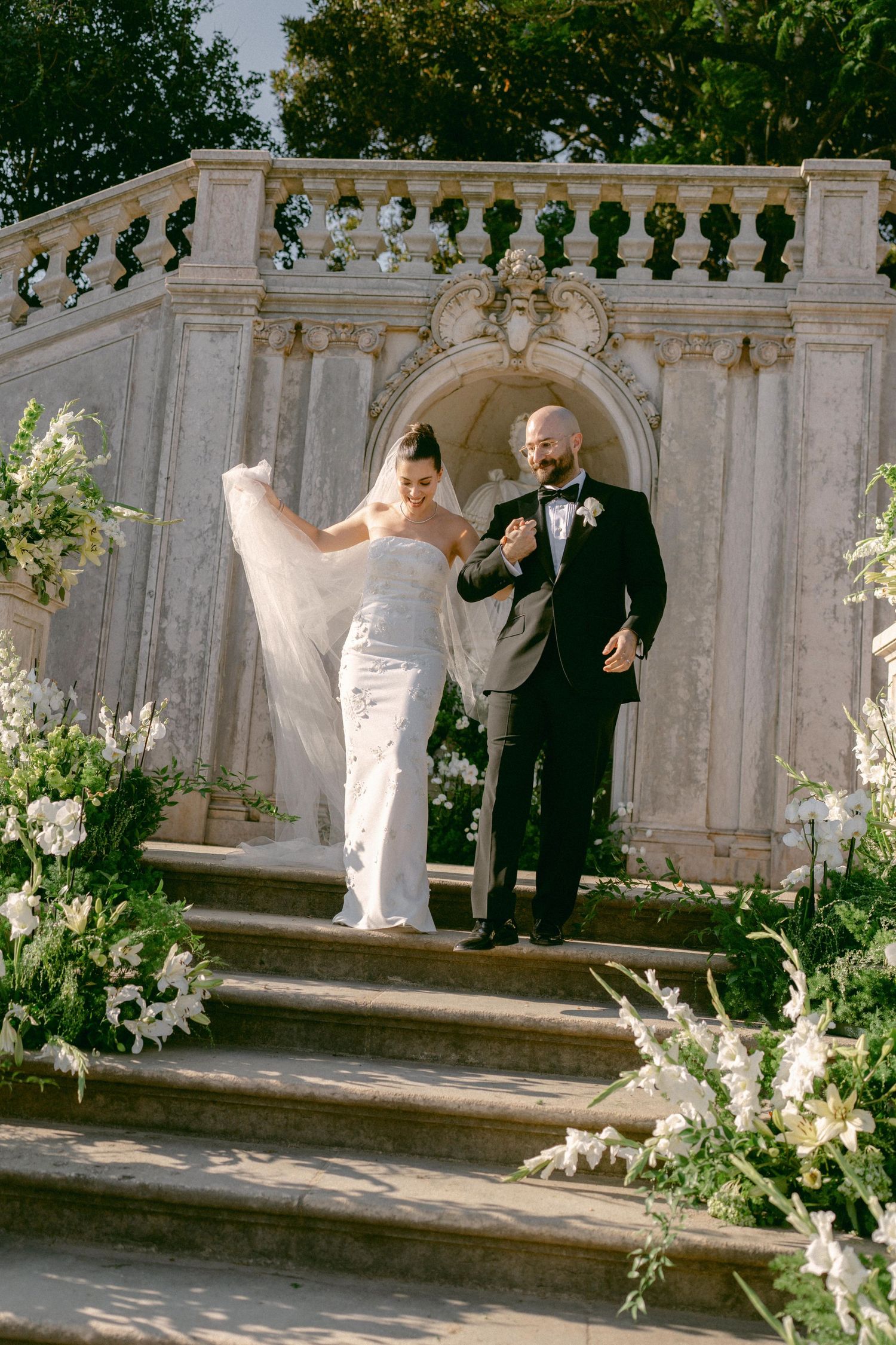 Bride and groom descending ornate stone staircase adorned with white florals after wedding ceremony at Vandelli Botanical Garden in Lisbon