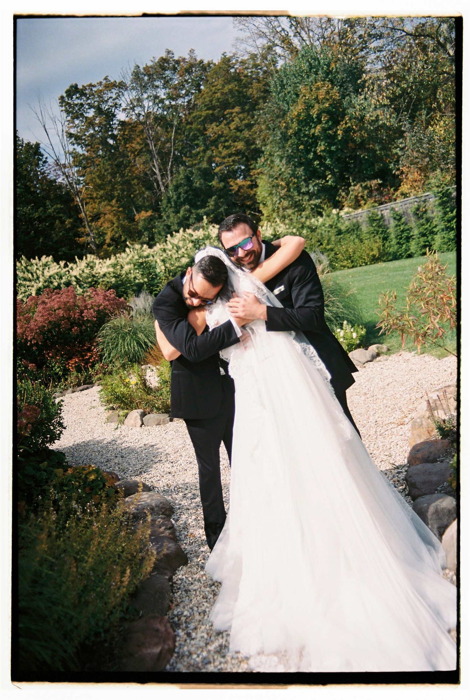 Bride and groom embracing and laughing in a garden setting, bride in white gown with veil, groom in black suit.