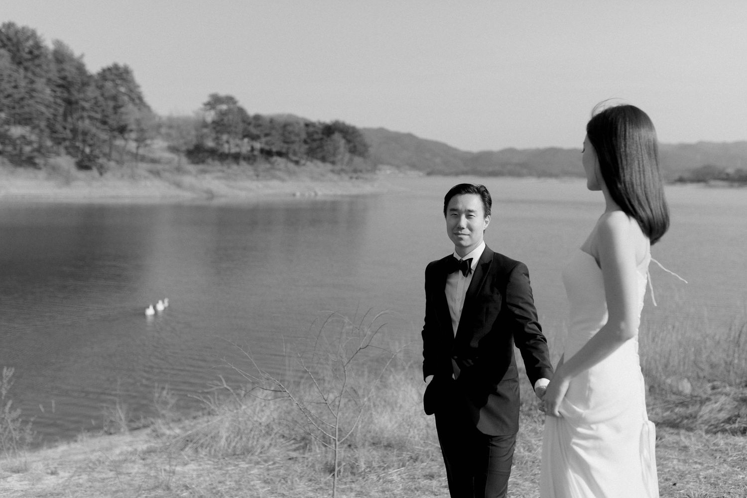 Couple sharing first look moment by lake with swans, groom in tuxedo