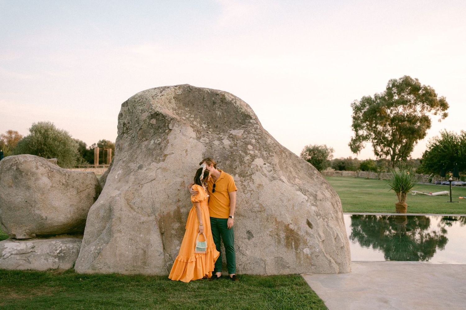 Bride and groom embracing near large boulders by reflective pool at sunset during rehearsal dinner at São Lourenço do Barrocal in Monsaraz