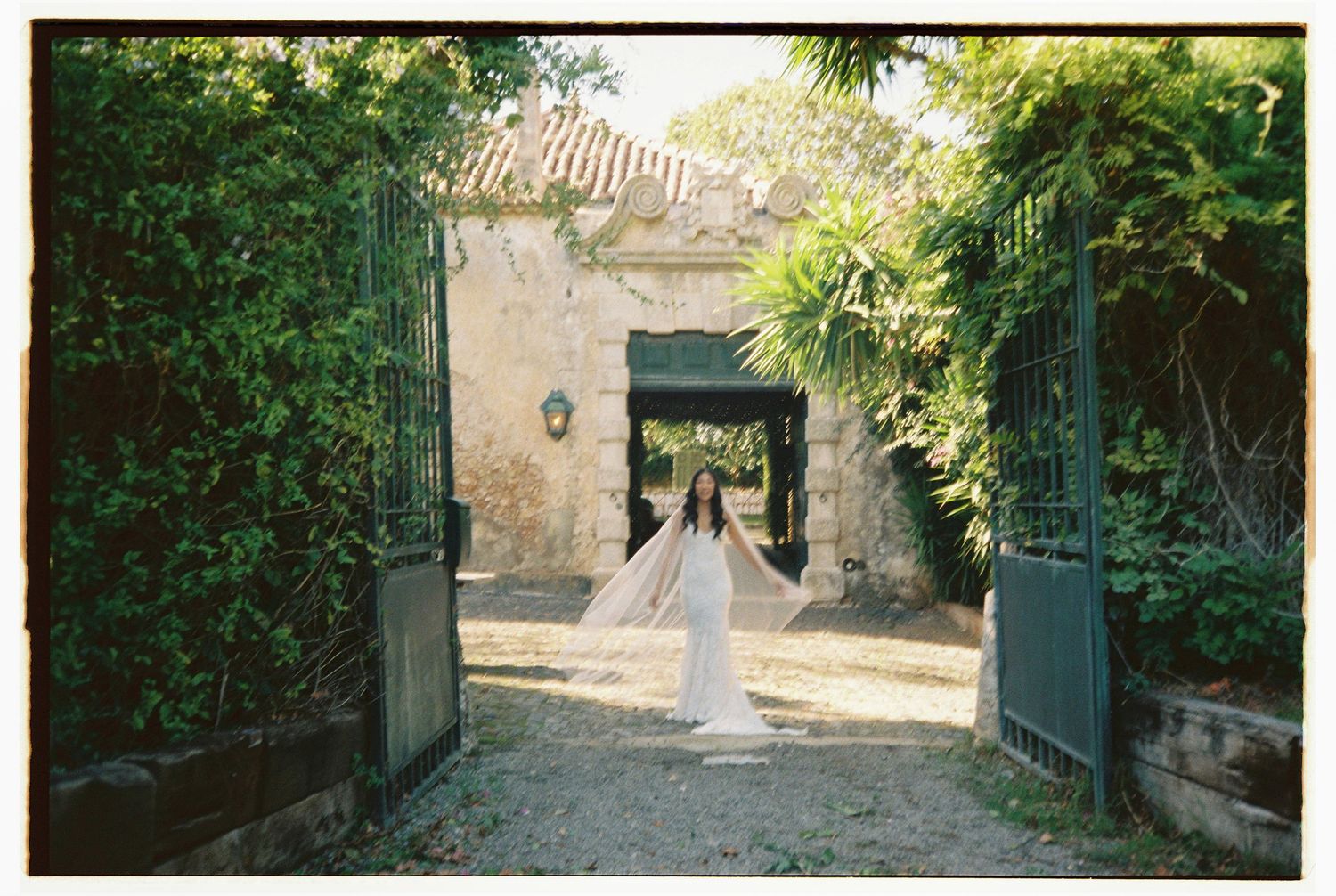Bride in white gown and veil standing between iron gates of stone estate
