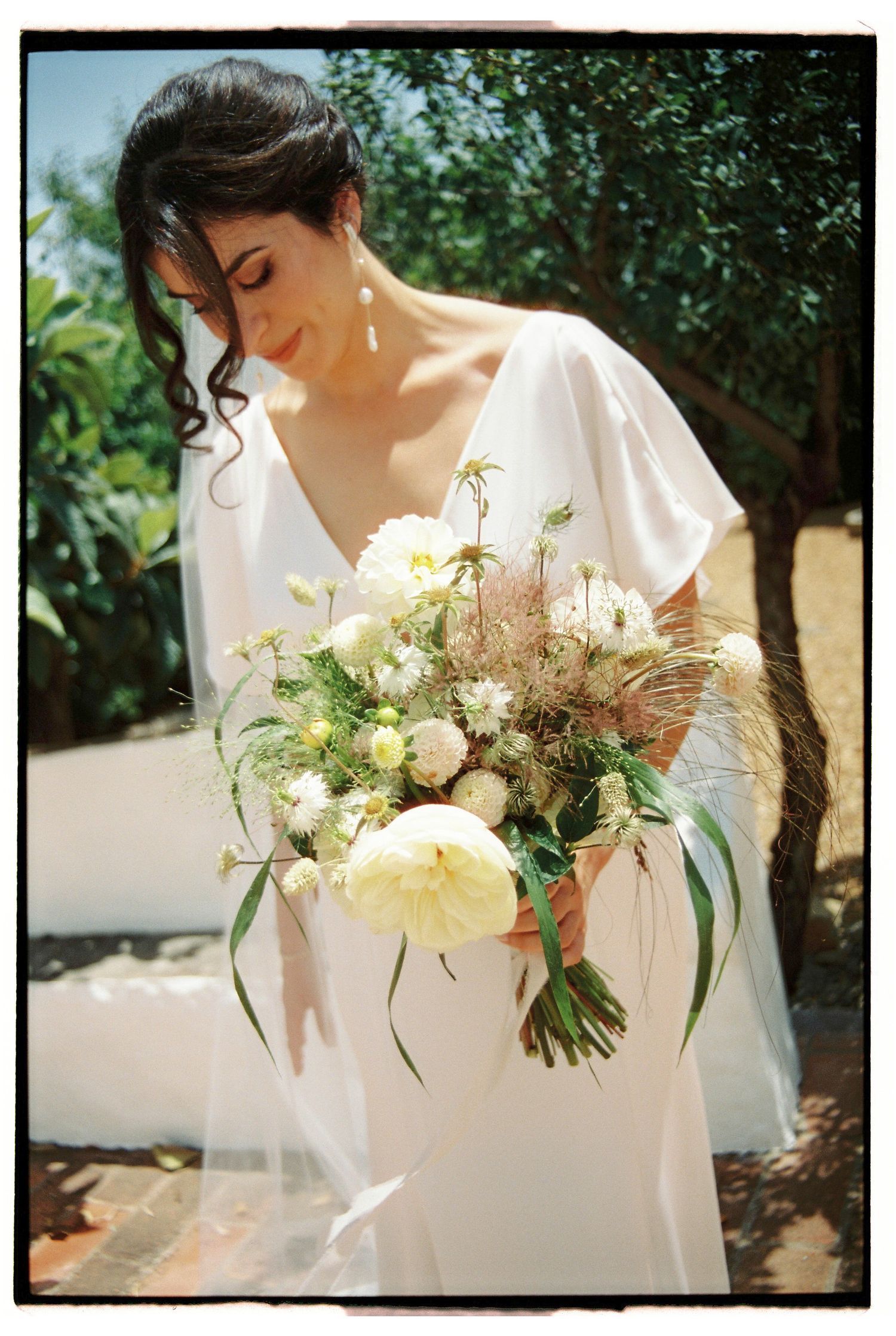 Bride holding white and green bouquet outdoors near olive trees during wedding