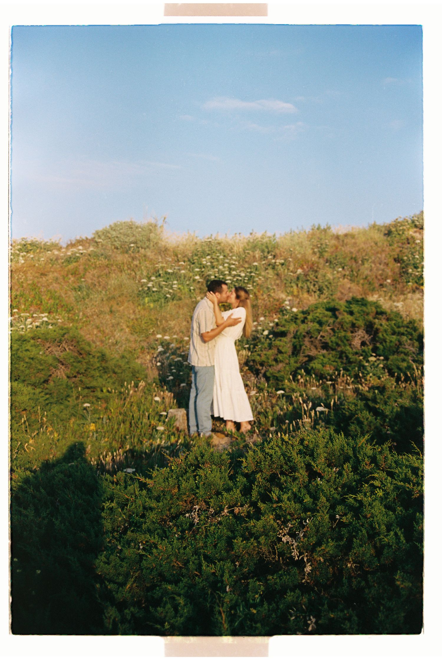 Couple sharing a kiss on a wildflower hillside under a bright blue sky on a film photo.