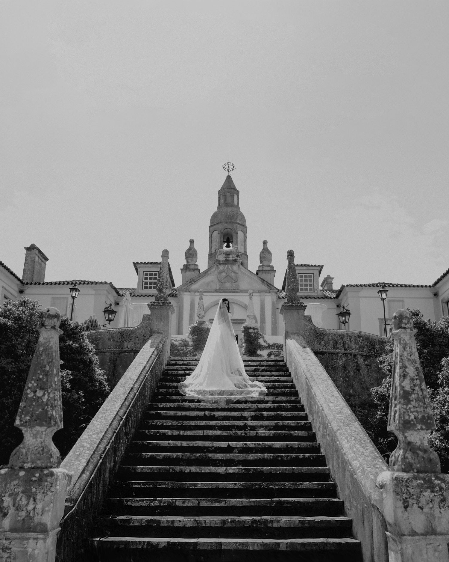 Bride in white gown and veil standing at base of grand stone staircase leading to historic venue at Quinta da Conceição in Azeitão