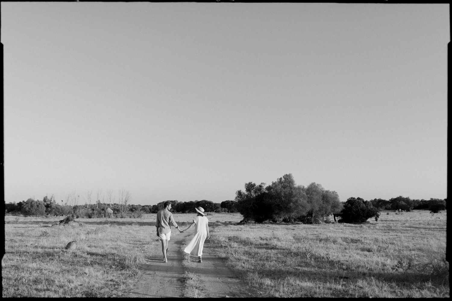 Couple in white attire walking along rural dirt path through open countryside fields