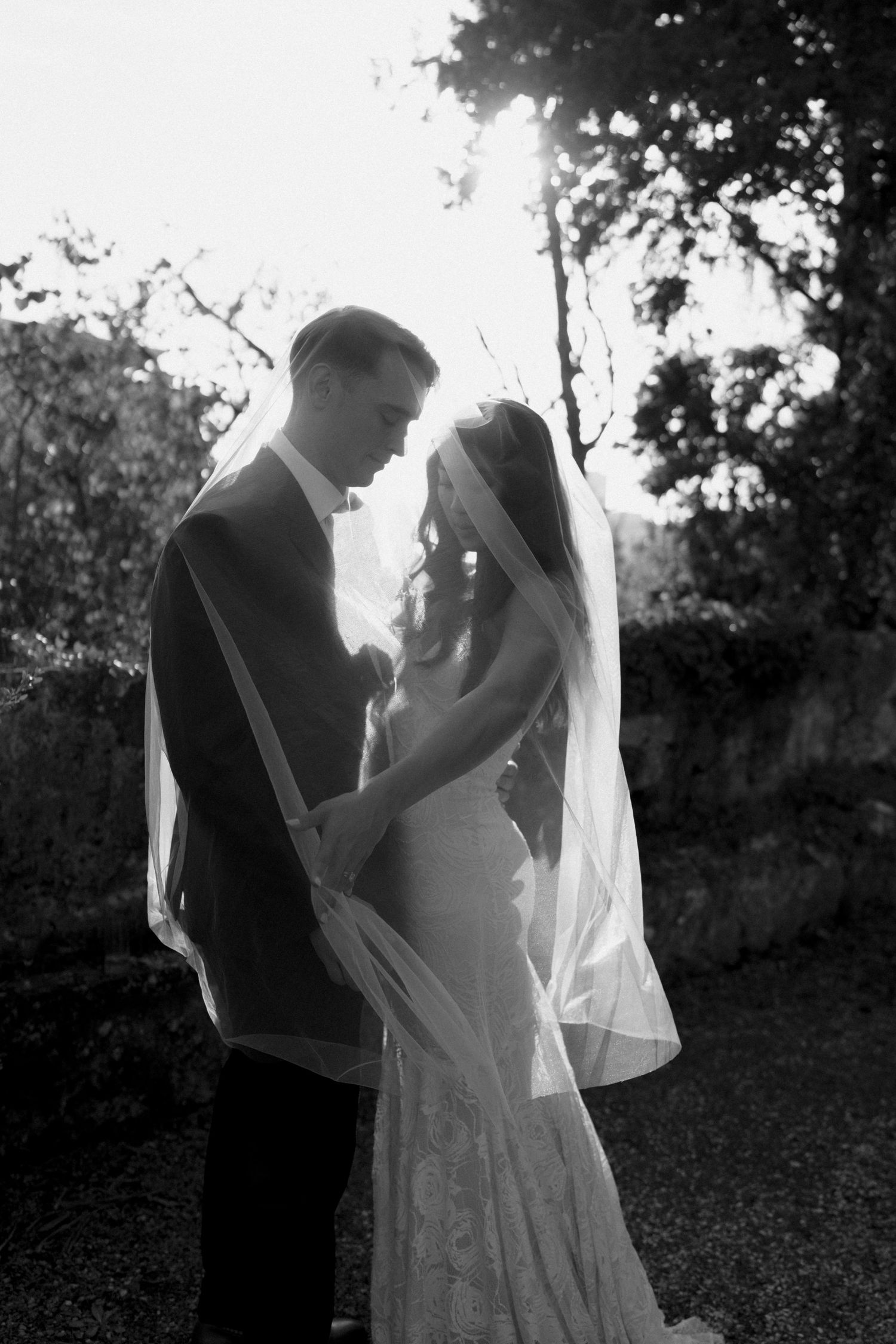 Backlit black and white portrait of bride and groom embracing under wedding veil outdoors