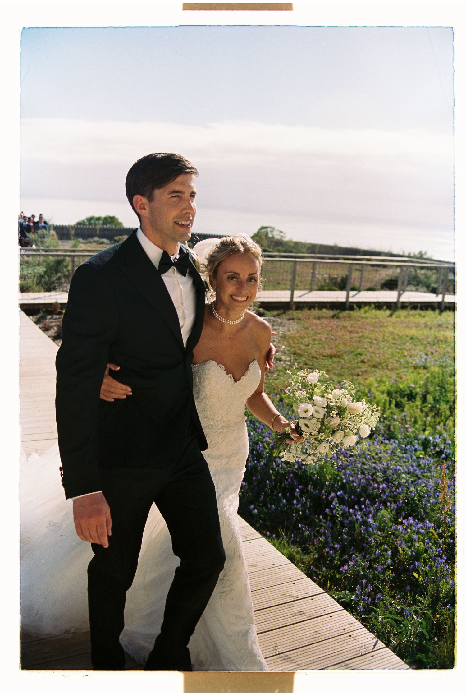 Bride and groom walking on wooden boardwalk surrounded by wildflowers during wedding at Aethos Ericeira