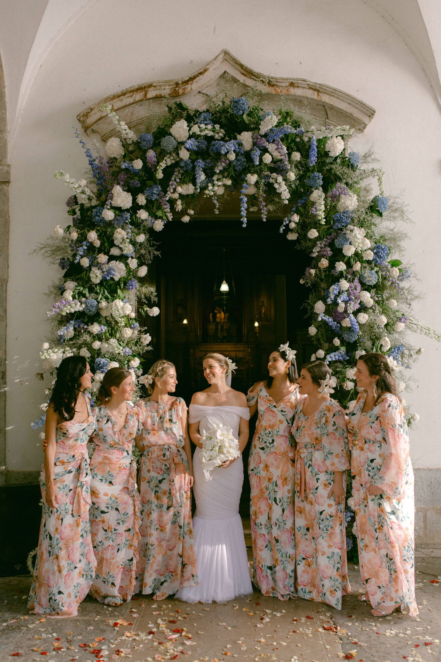 Bride with bridesmaids in floral dresses standing under blue and white floral arch doorway