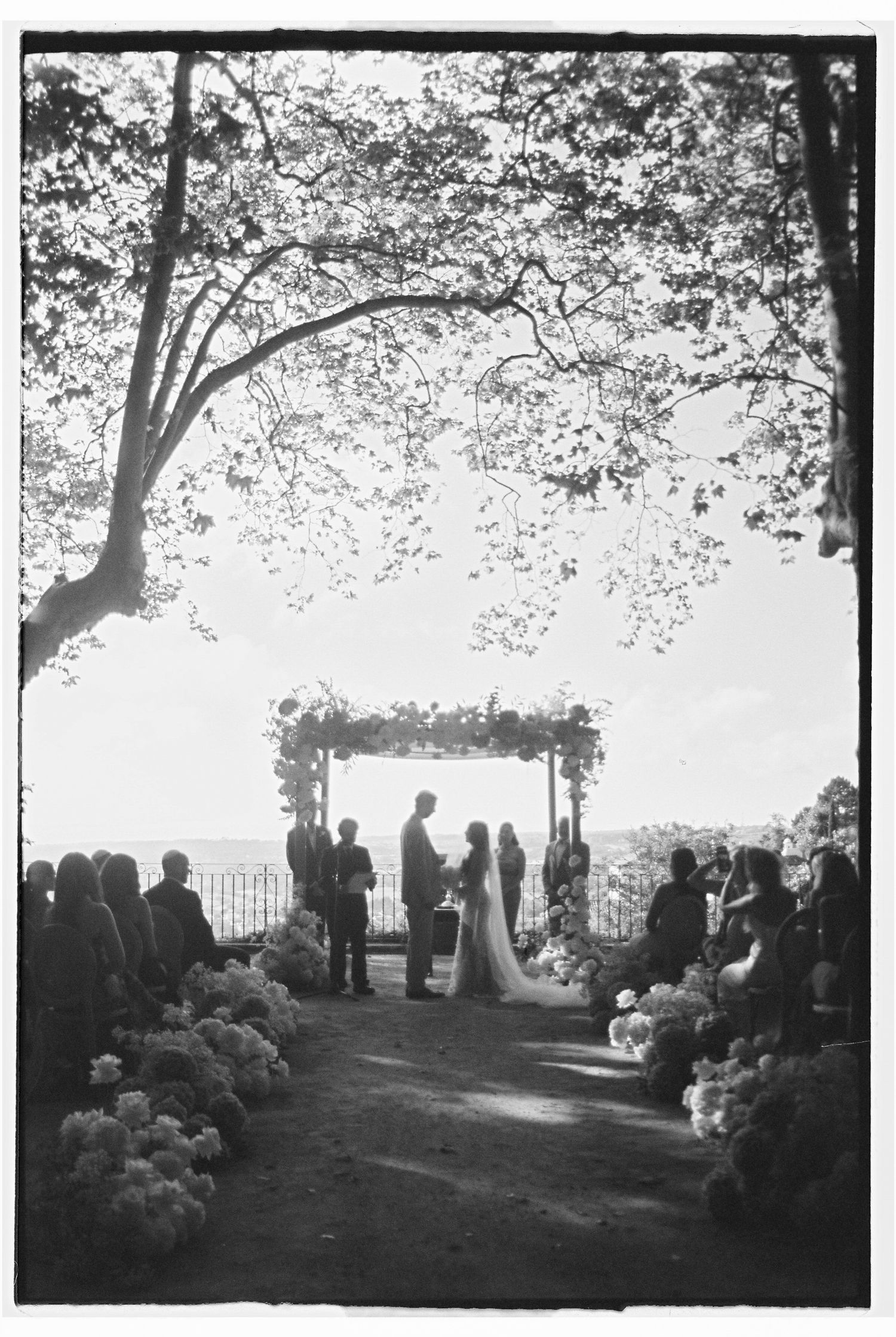 Outdoor wedding ceremony under floral arch framed by trees with guests and floral aisle décor, backlit at Quinta da Bellavista in Sintra