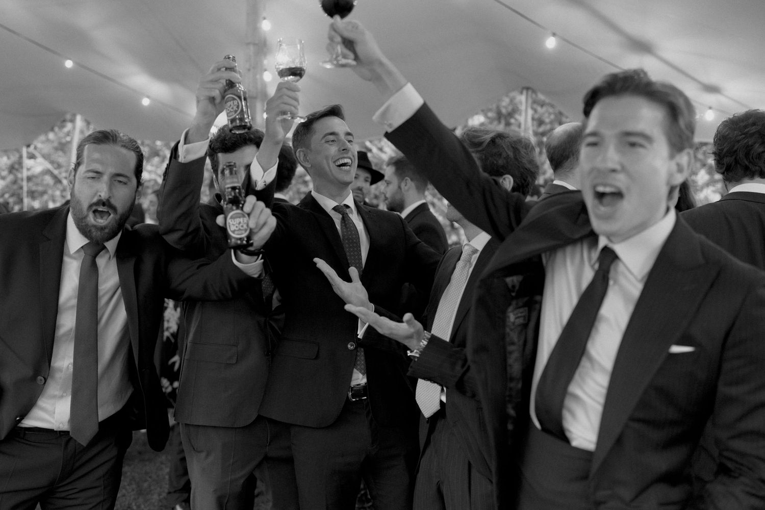 Groomsmen celebrate joyfully raising drinks in black and white photo at wedding reception tent.