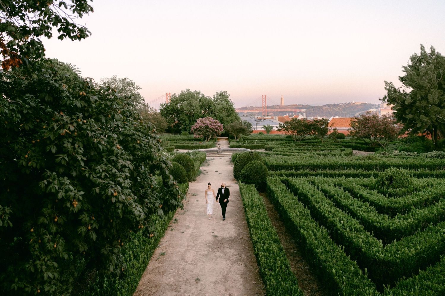 Person walking through a formal hedge maze garden with a bridge and city skyline at dusk.