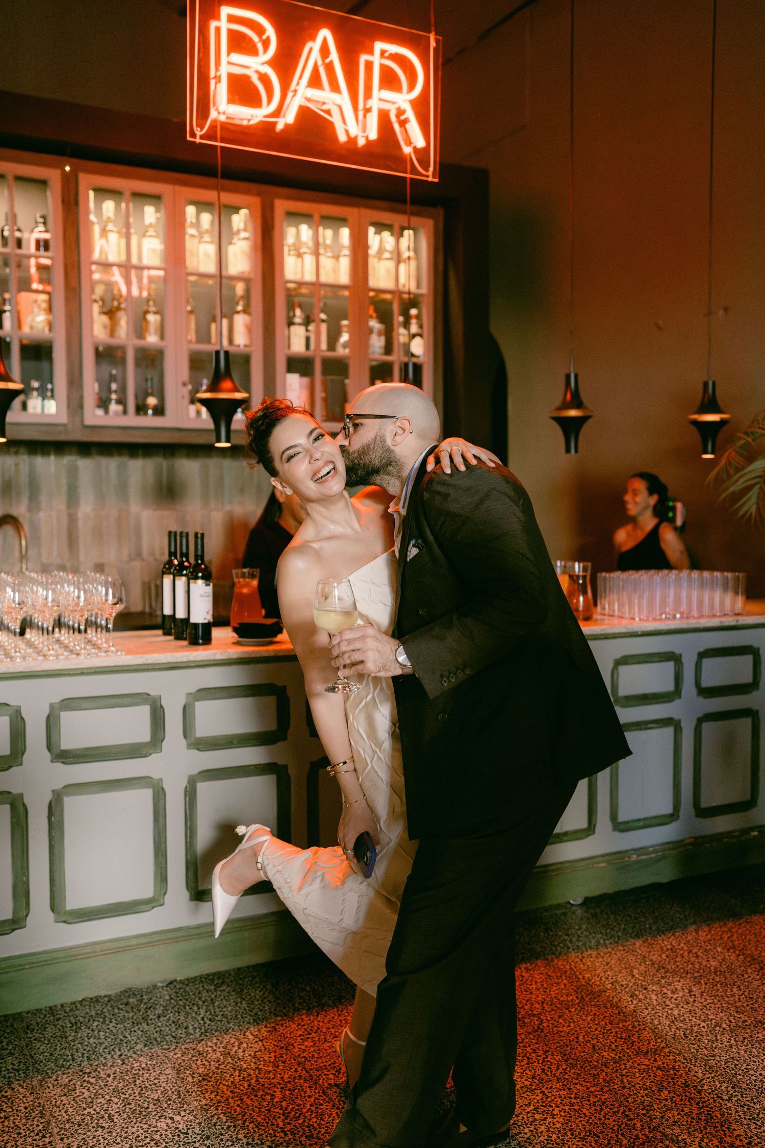 A joyful couple laughs and embraces in front of a glowing neon BAR sign at their wedding reception.