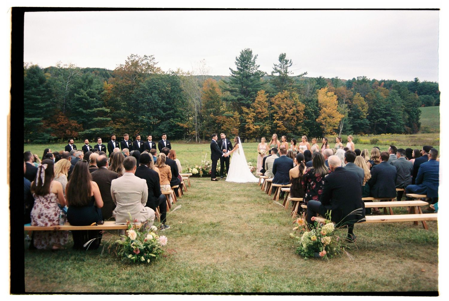 Outdoor wedding ceremony on green lawn with guests seated on wooden benches surrounded by autumn trees at Hayfield in Catskills, New York