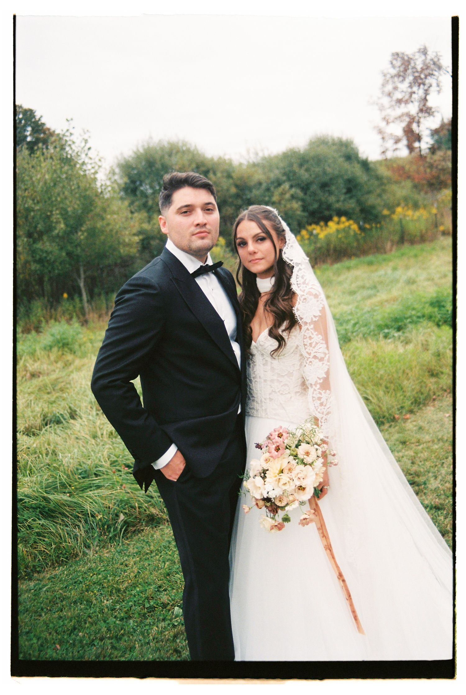 Bride and groom pose outdoors in elegant wedding attire, she holds a pastel bouquet with a long veil.