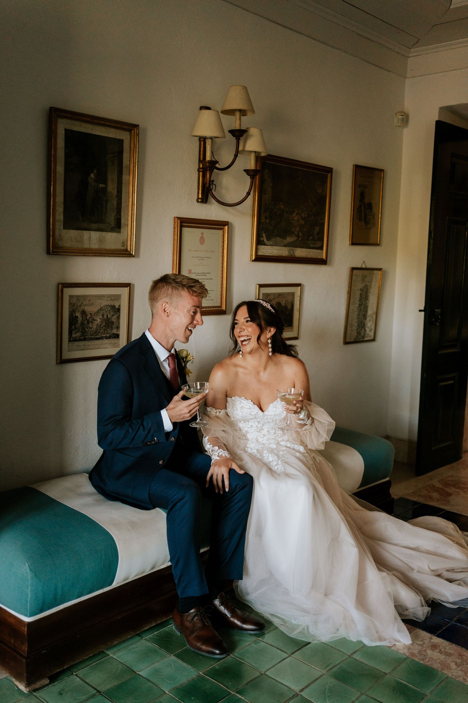 Bride and groom sitting together on a teal bench, laughing and toasting with champagne in an elegant room with framed artwork.