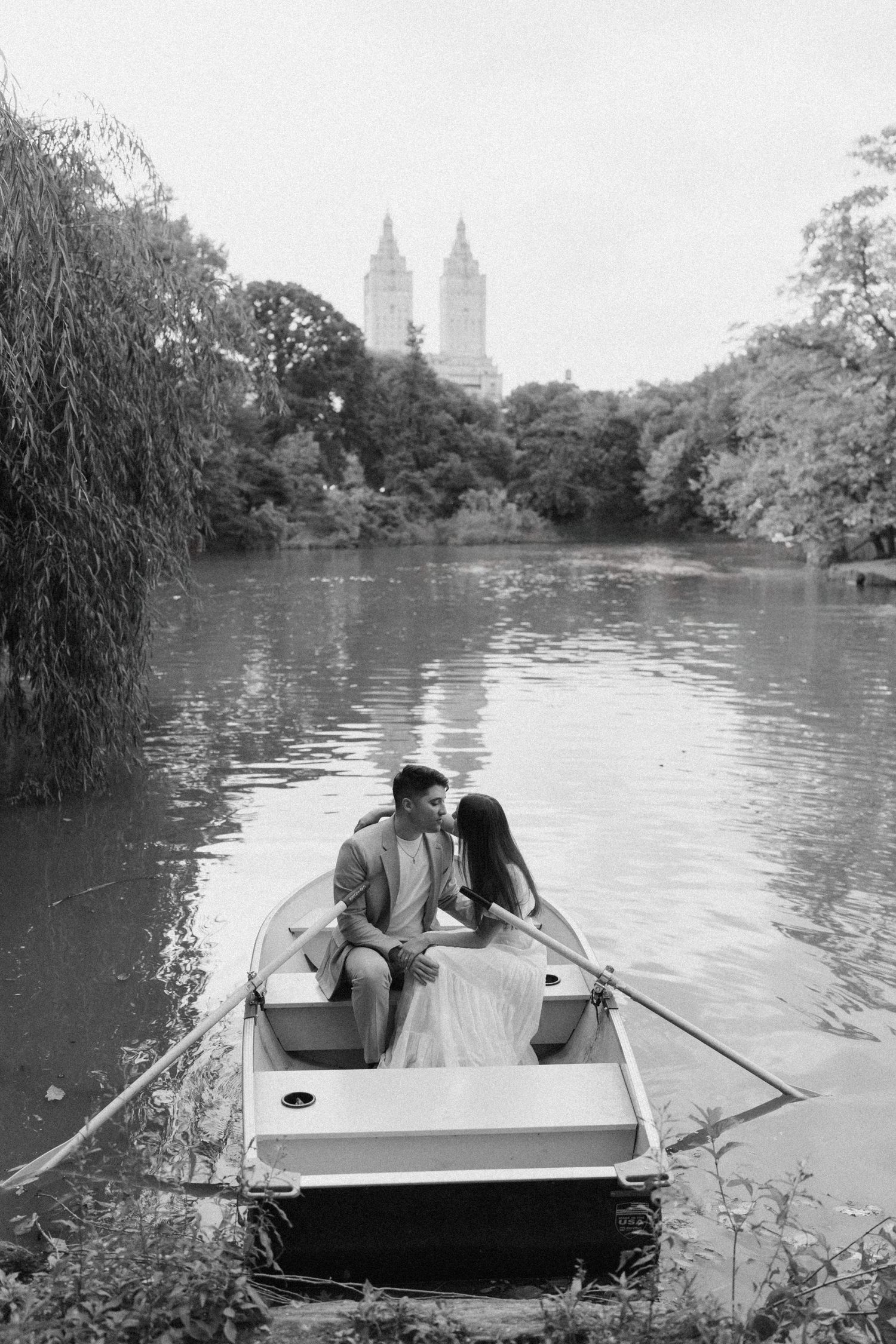 Black and white photo of couple sharing intimate moment in rowboat on Central Park lake with skyline.