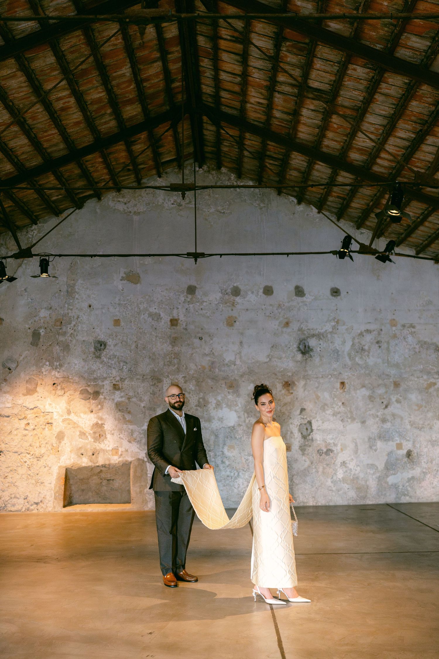 Bride in white gown and groom in dark suit pose in rustic stone venue with wooden ceiling.