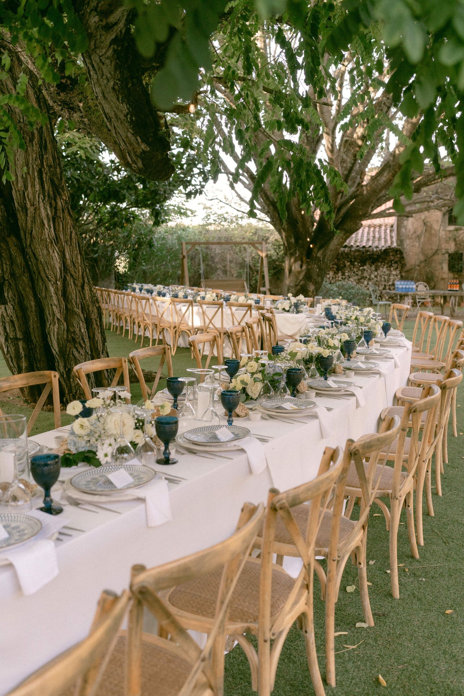 Outdoor wedding reception with long white tables, wooden chairs, blue glassware, and white floral centerpieces under trees