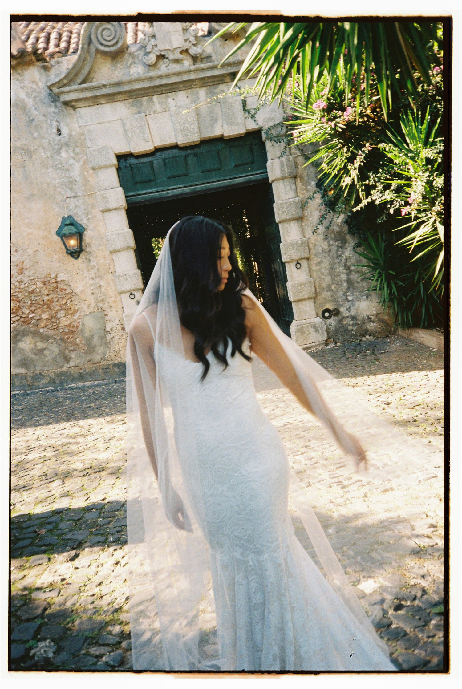 Bride in lace wedding dress and veil twirling outside rustic stone building