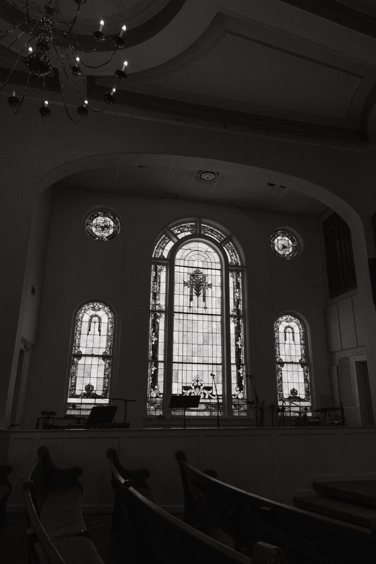Black and white photo of church interior with ornate stained glass windows and wooden pews under a chandelier.