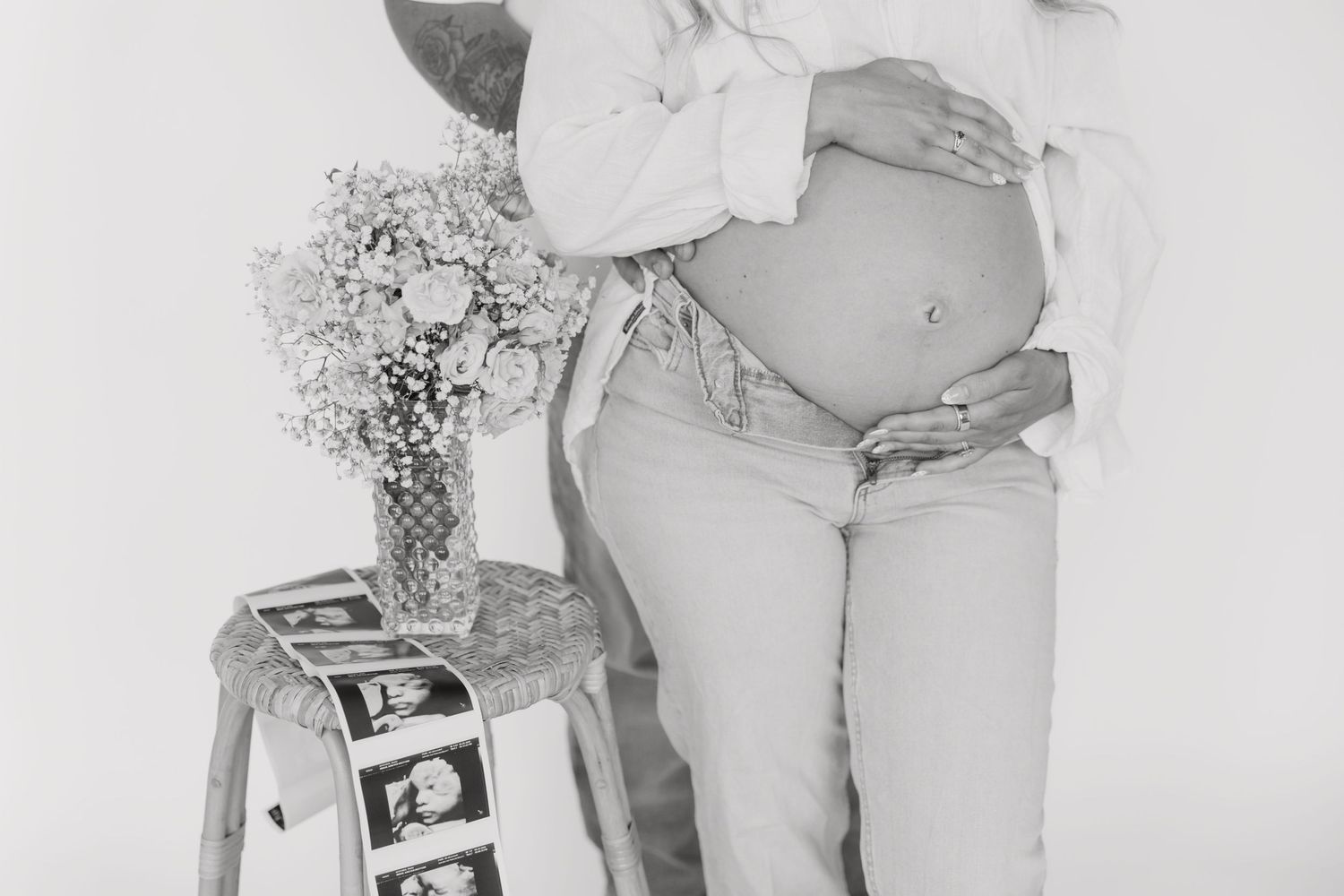 Black and white maternity photo of pregnant woman holding belly with ultrasound photos and flowers on stool.
