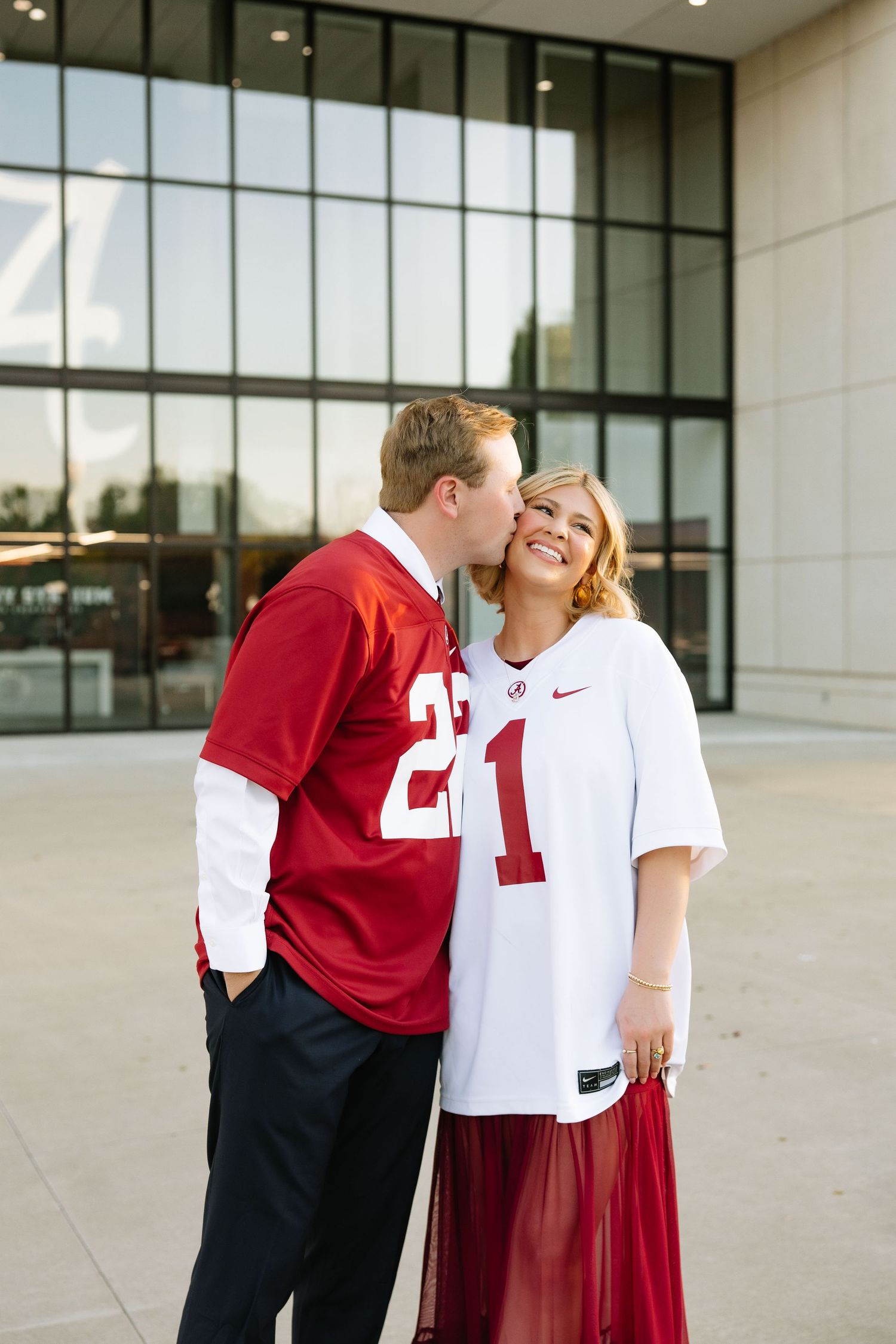 Couple in Alabama Crimson Tide jerseys sharing a kiss outside a stadium, wearing numbers 22 and 1.