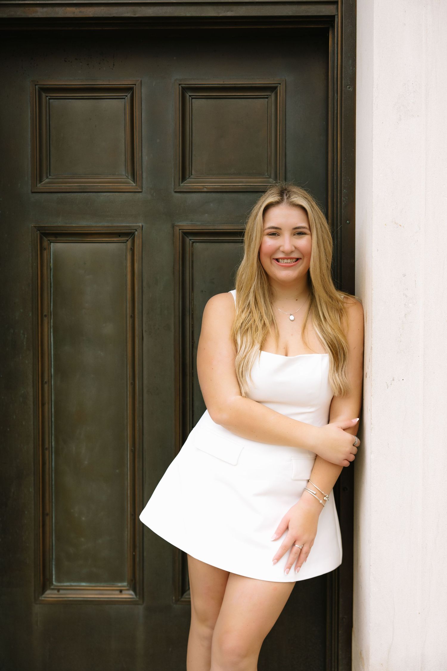 Smiling young woman in white strapless mini dress leaning against a dark door outdoors.