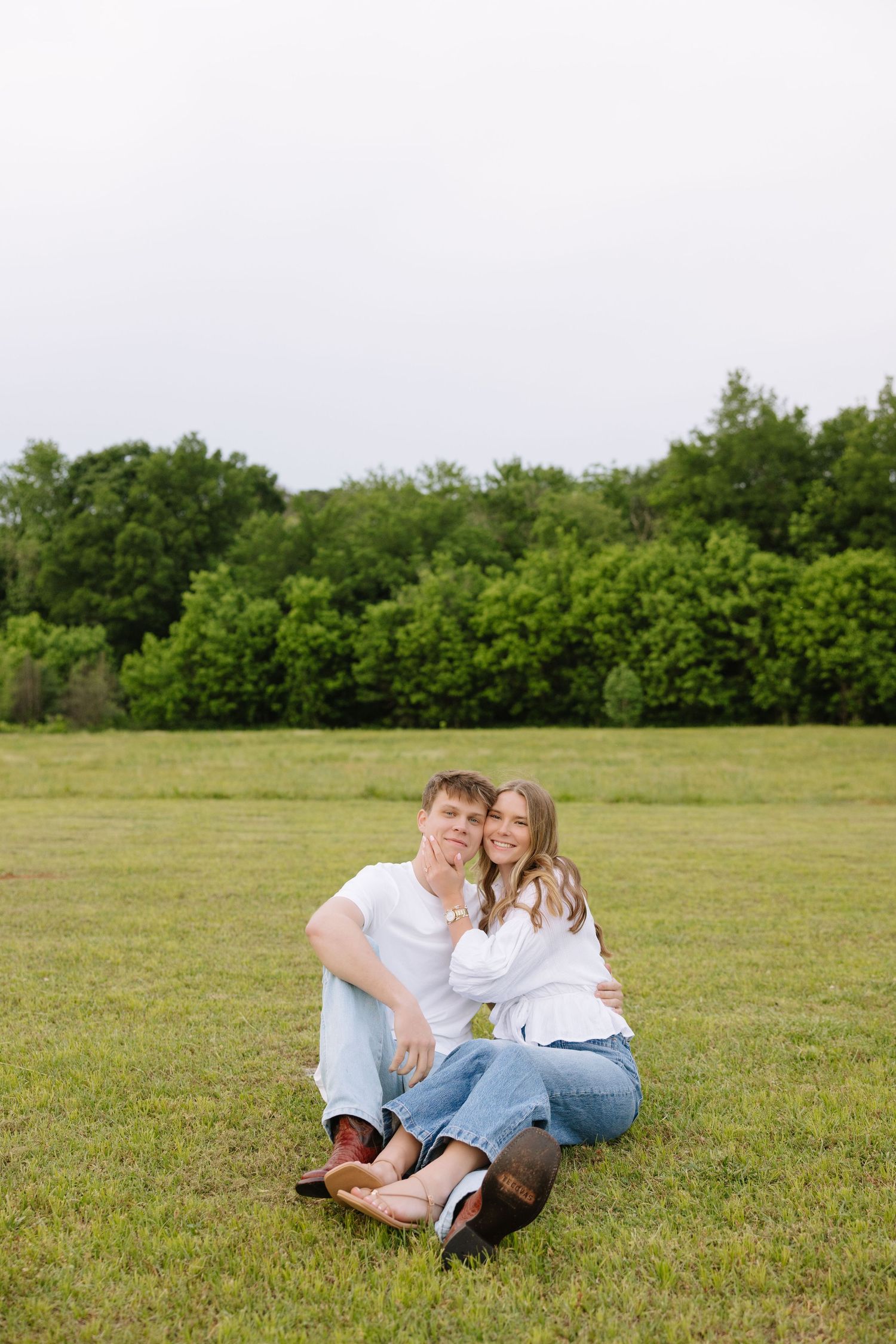 Happy couple in white shirts and jeans sitting together on green grass field with trees in background.