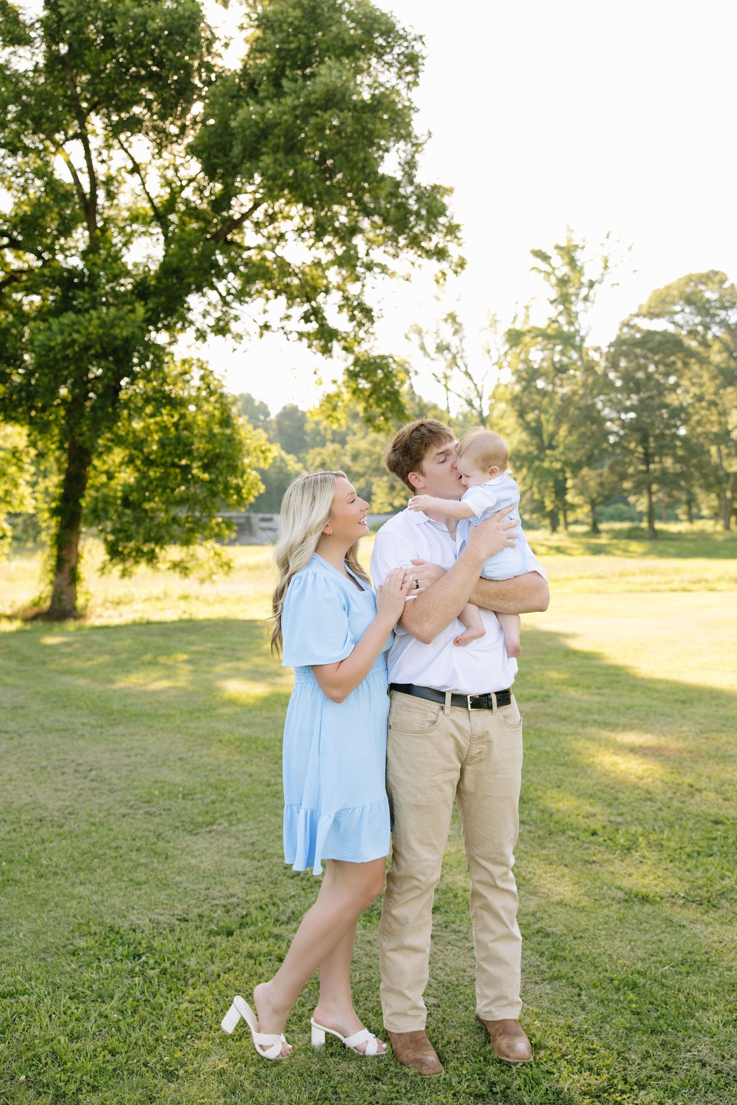 Young family with baby poses outdoors in sunny green park, mom in blue dress and dad in white shirt holding infant.